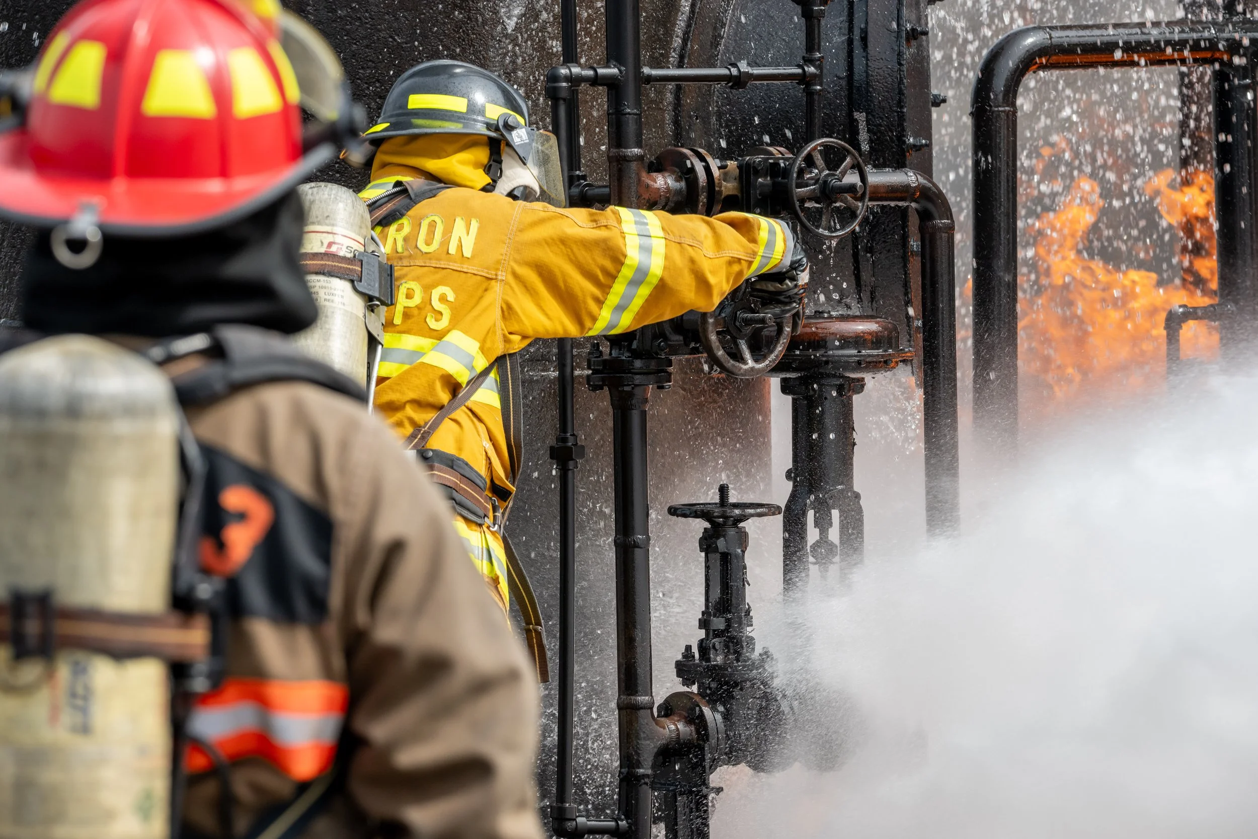 Firefighters in protective gear fighting a fire with water and steam, with flames visible in the background.