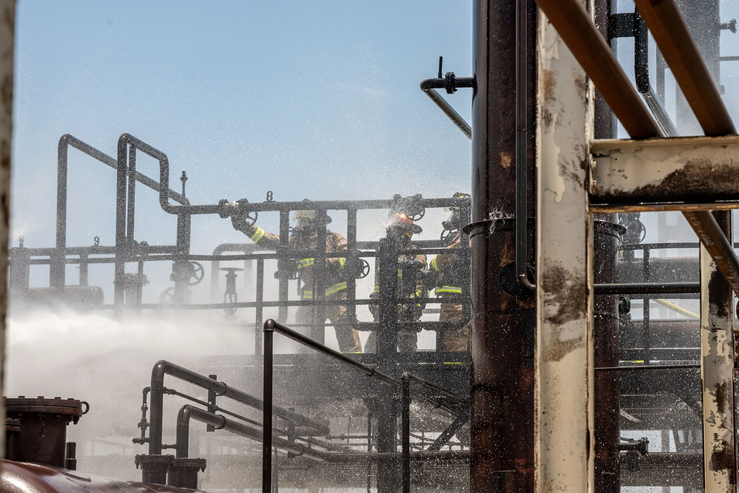 Firefighters spray water on a burning industrial structure with rusted pipes and metal scaffolding.