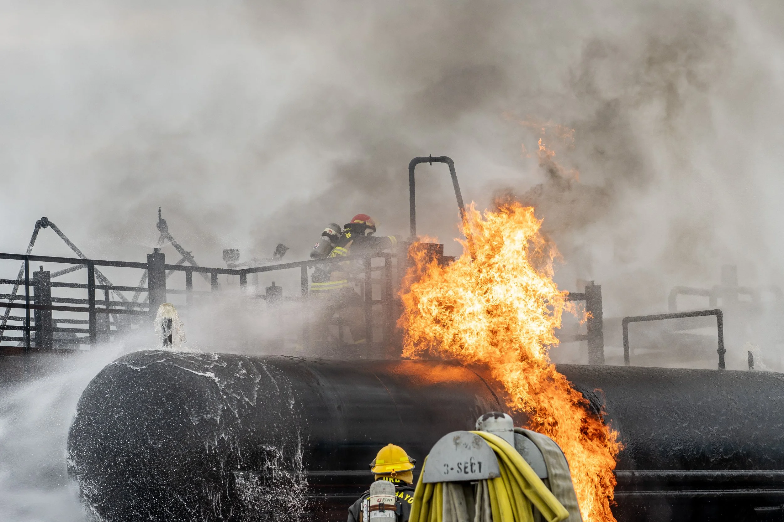 Firefighters battling a large fire on an industrial tank with flames and thick smoke.