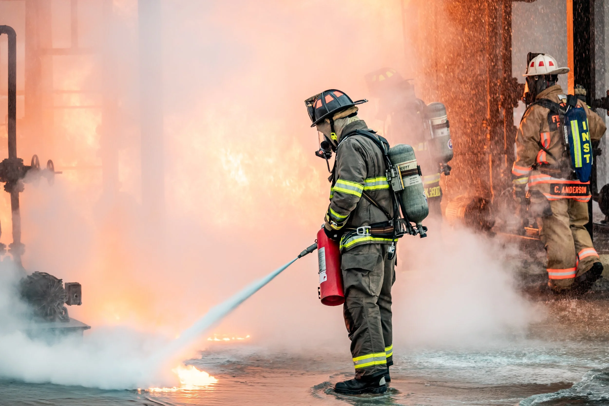 Firefighters in protective gear combat a blaze, one spraying water and others managing the flames at the scene of a fire.