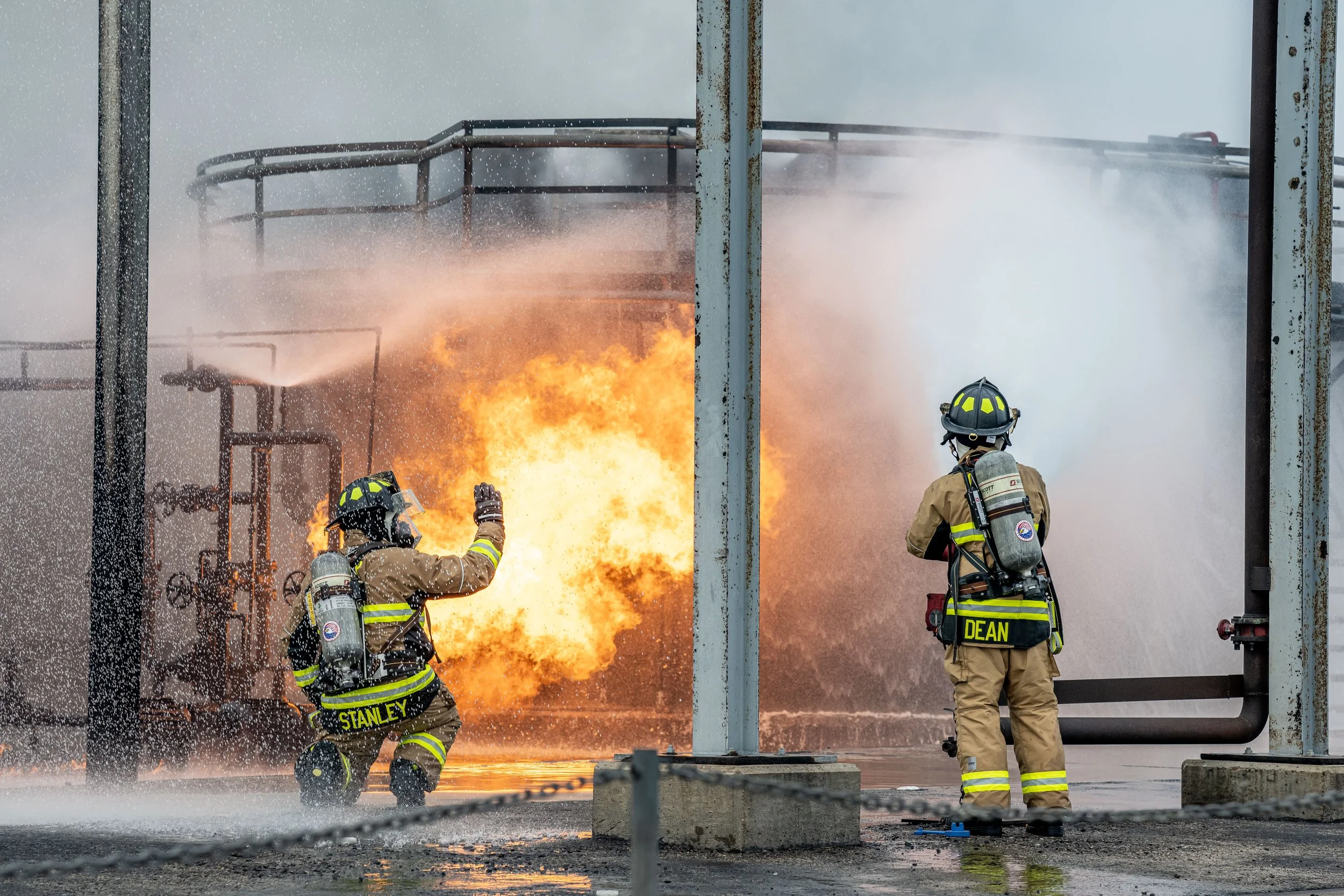 Two firefighters, one kneeling and one standing, extinguish a large fire at an industrial site with flames and smoke in the background.