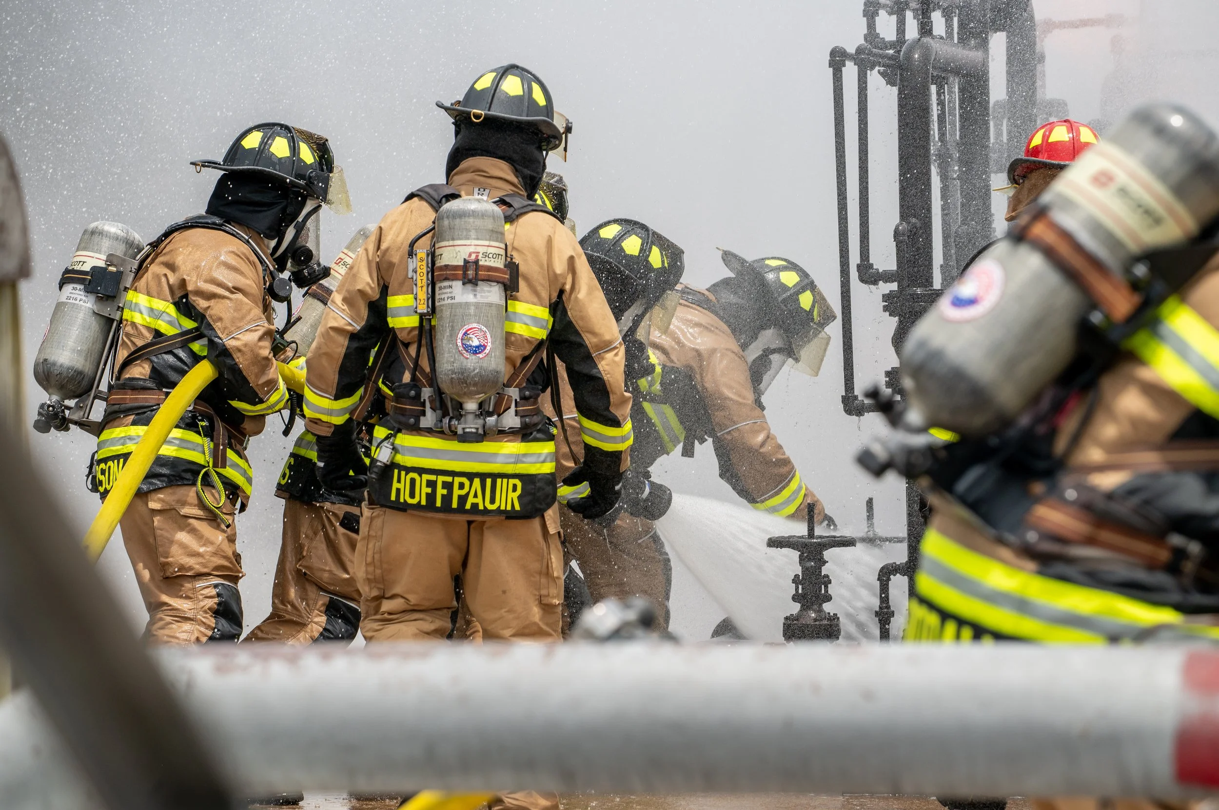 Group of firefighters in full gear battling a fire with water hose, wearing protective clothing and helmets, with water spraying and smoke in the background.