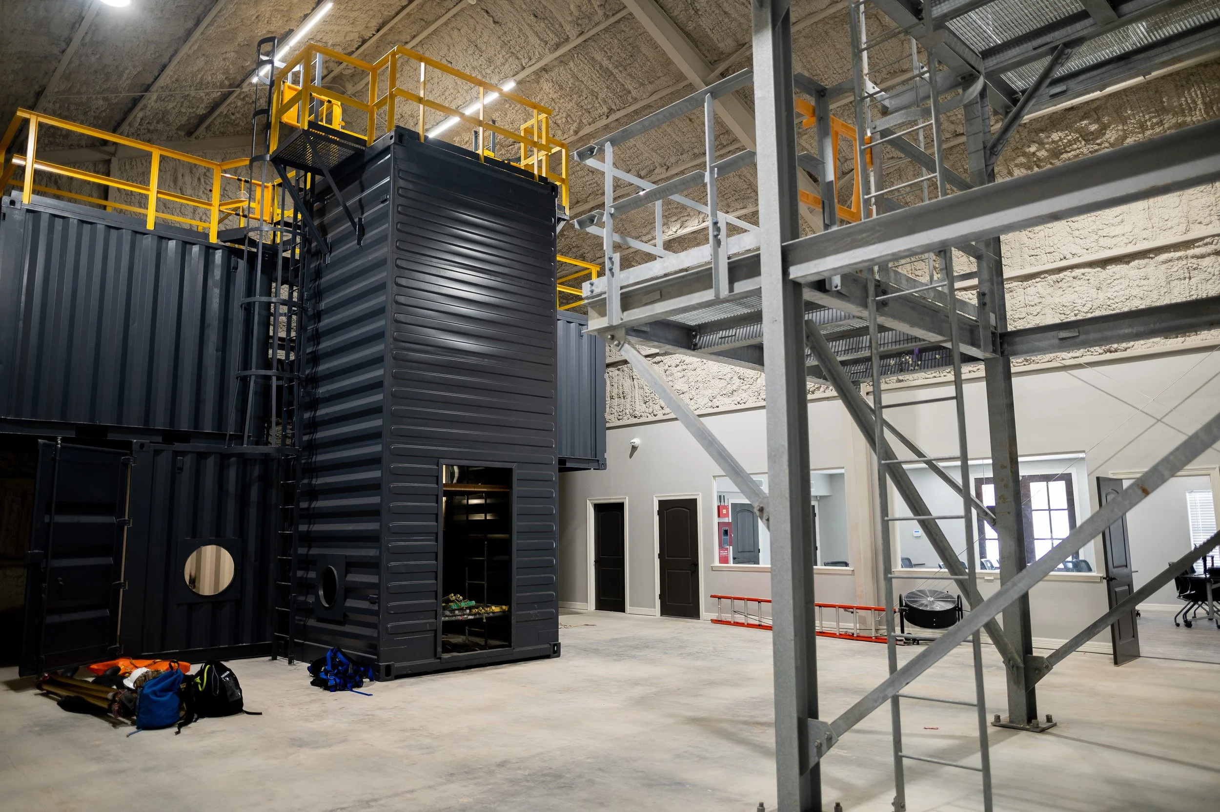 Interior of a building with black container structures, a metal scaffolding, and construction equipment, including backpacks and a ladder, indicating ongoing construction or renovation.