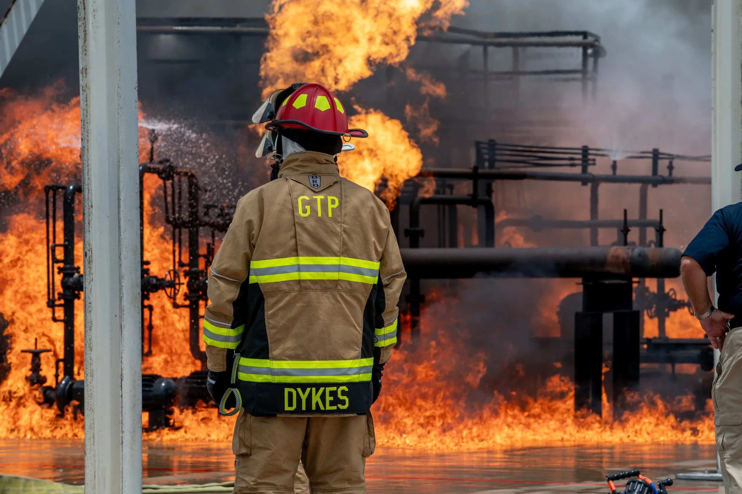 Firefighter in protective gear standing in front of a large fire at an industrial facility with flames and smoke.