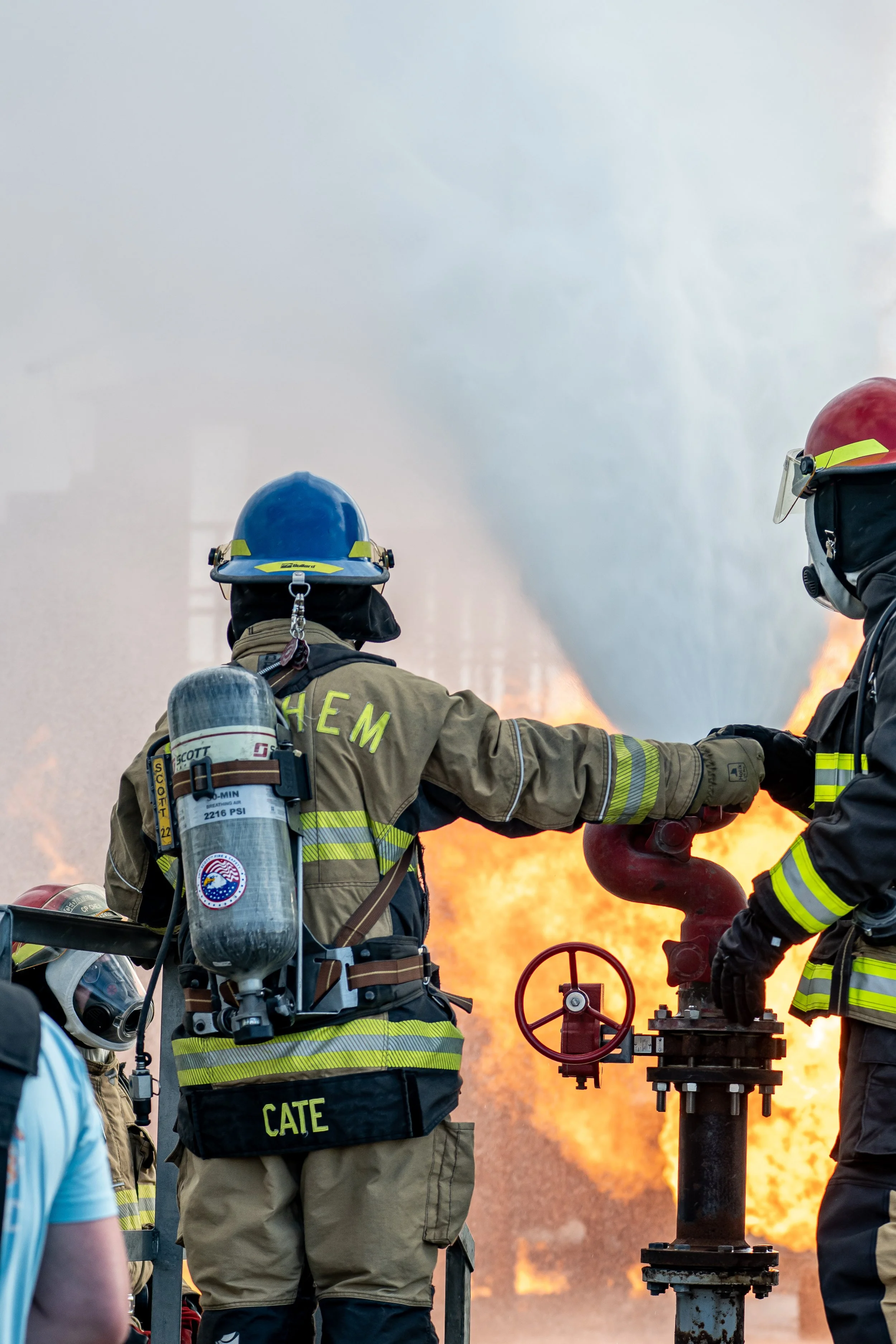 Firefighters in firefighting gear working at a fire scene with flames and smoke in the background.