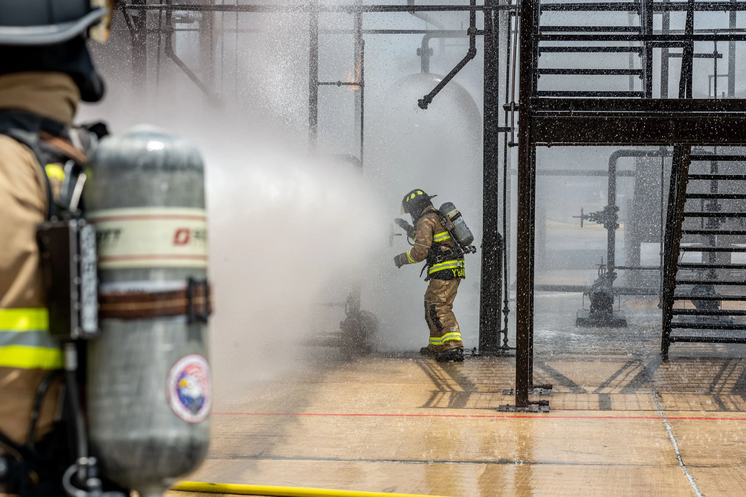 Firefighters in tan protective gear and helmets working to extinguish a fire, with one firefighter using a hose to spray water. Scaffolding and industrial pipes are visible in the background, and water spray creates a mist around the scene.