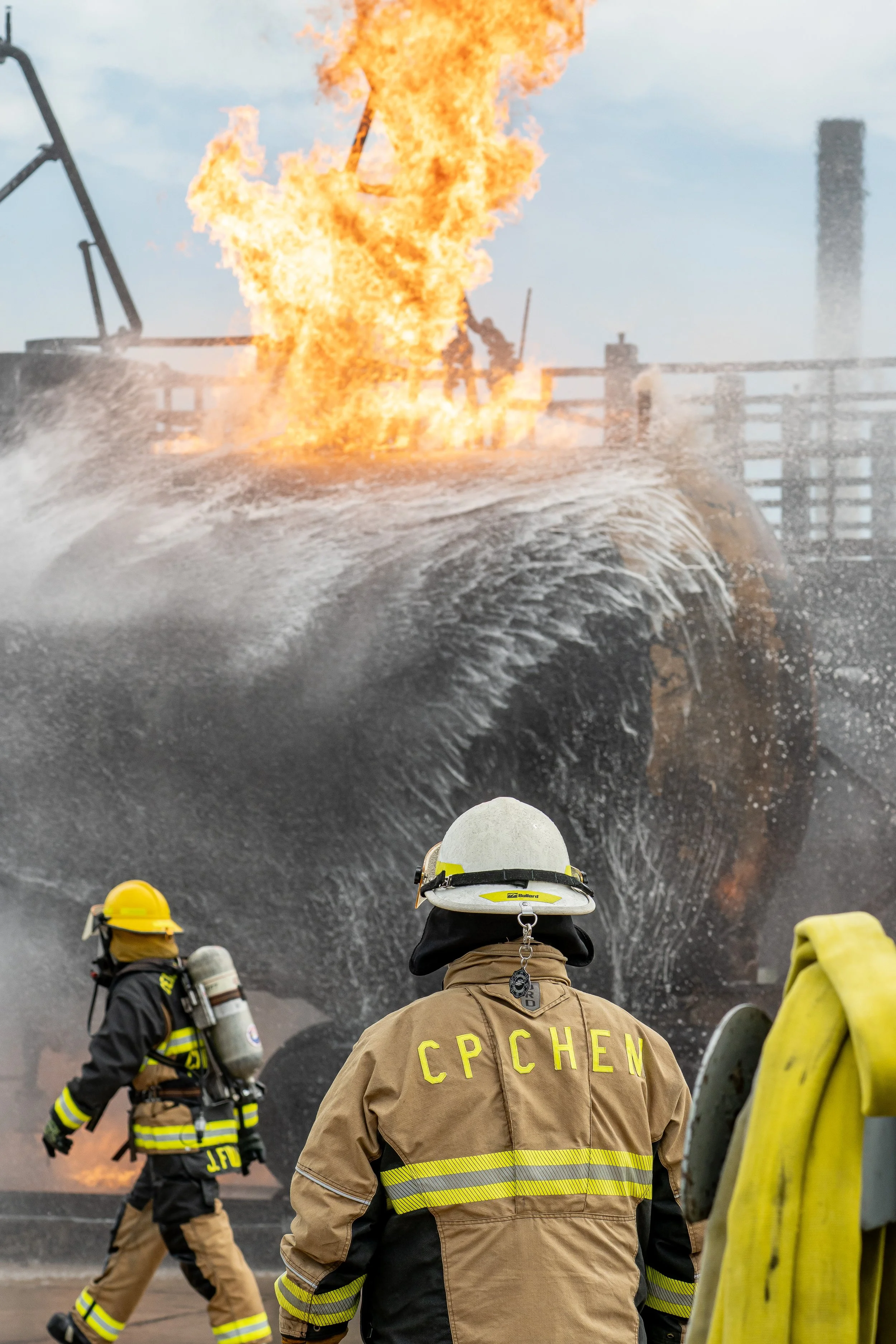 Firefighters in protective gear respond to a vessel on fire with flames and smoke, at sea or dockside.