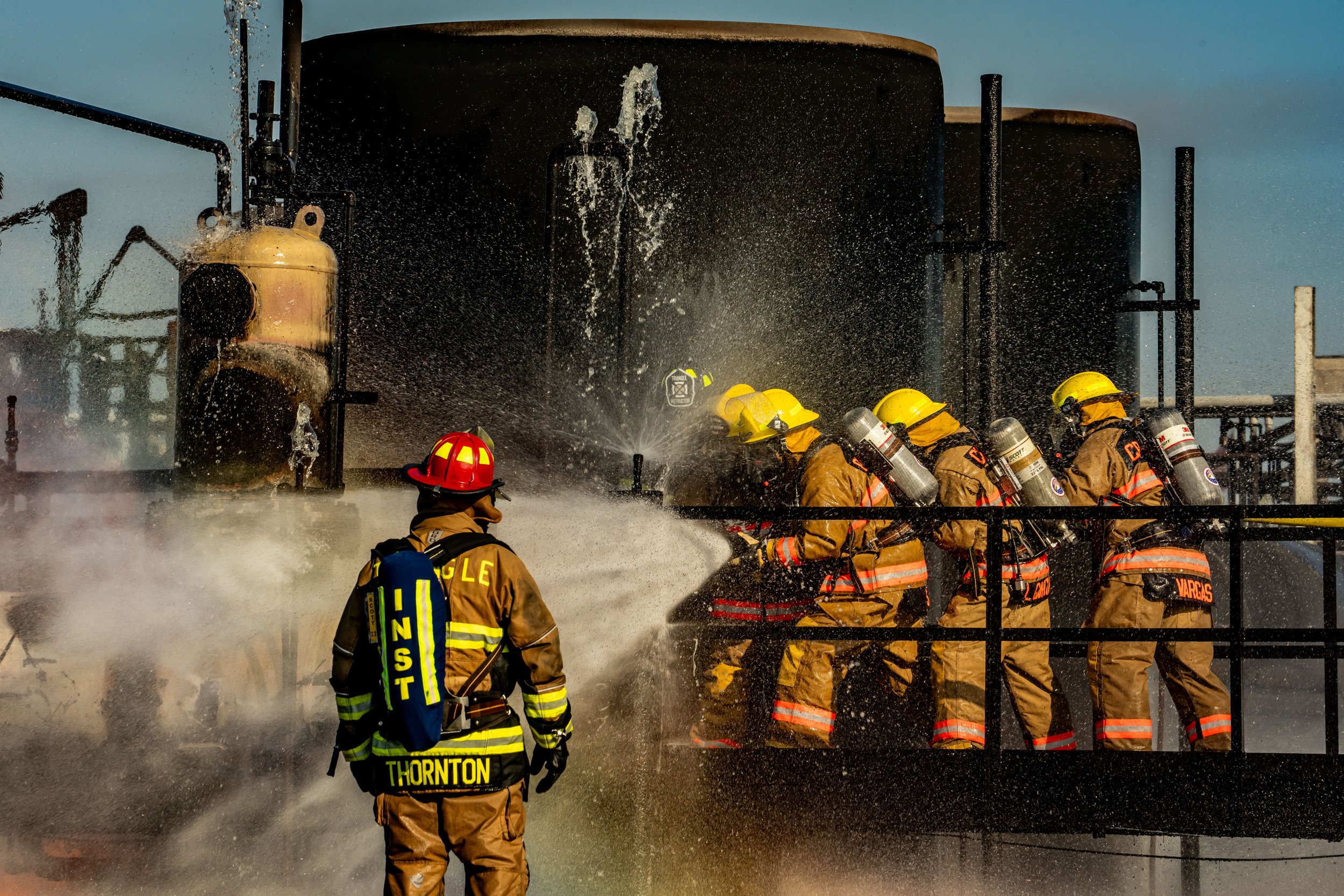 Firefighters in protective gear and helmets work together to extinguish a fire on industrial equipment at an industrial site, with water spray and steam in the air.