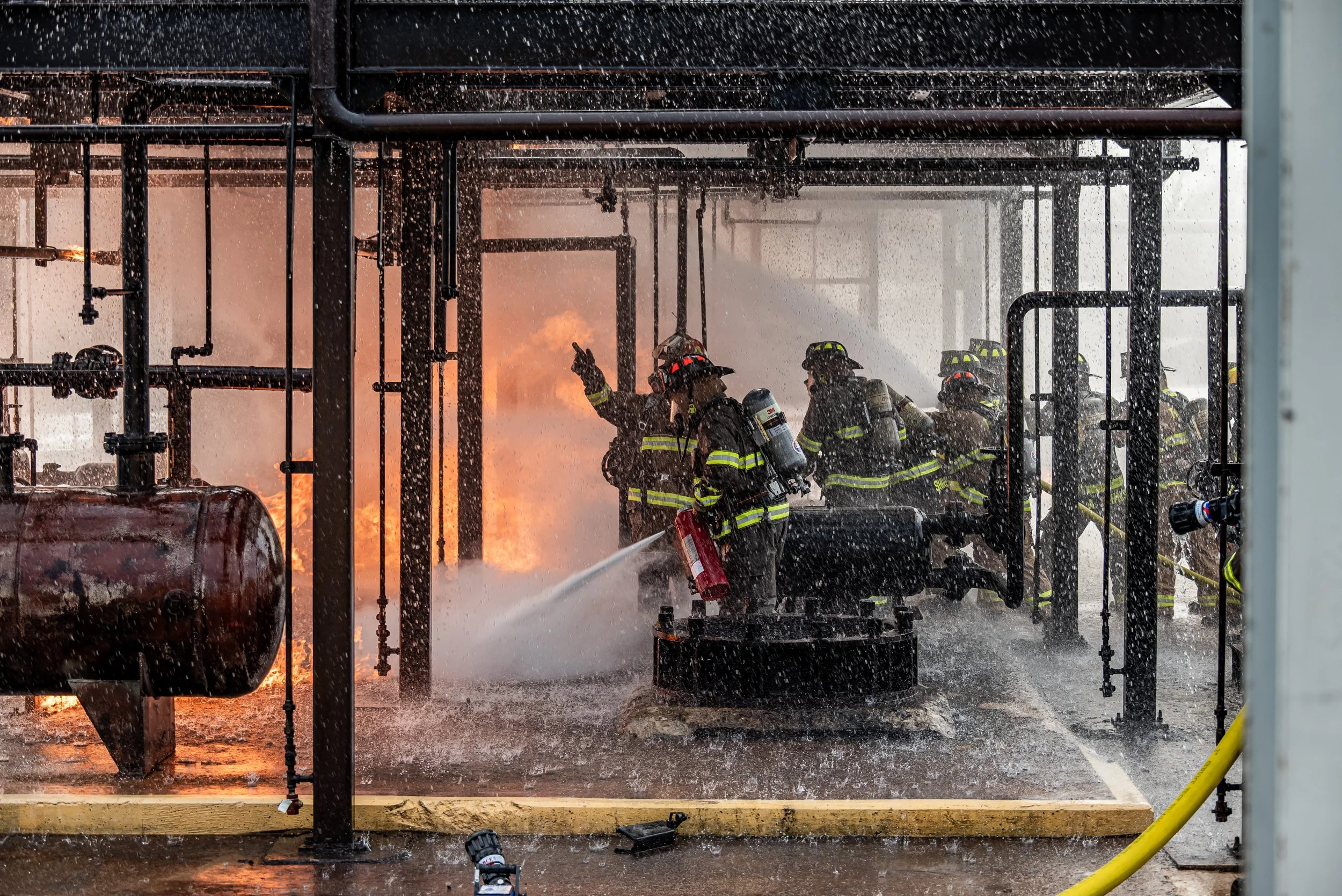 Firefighters extinguishing a fire with water spray in an industrial setting, flames and smoke visible in the background.