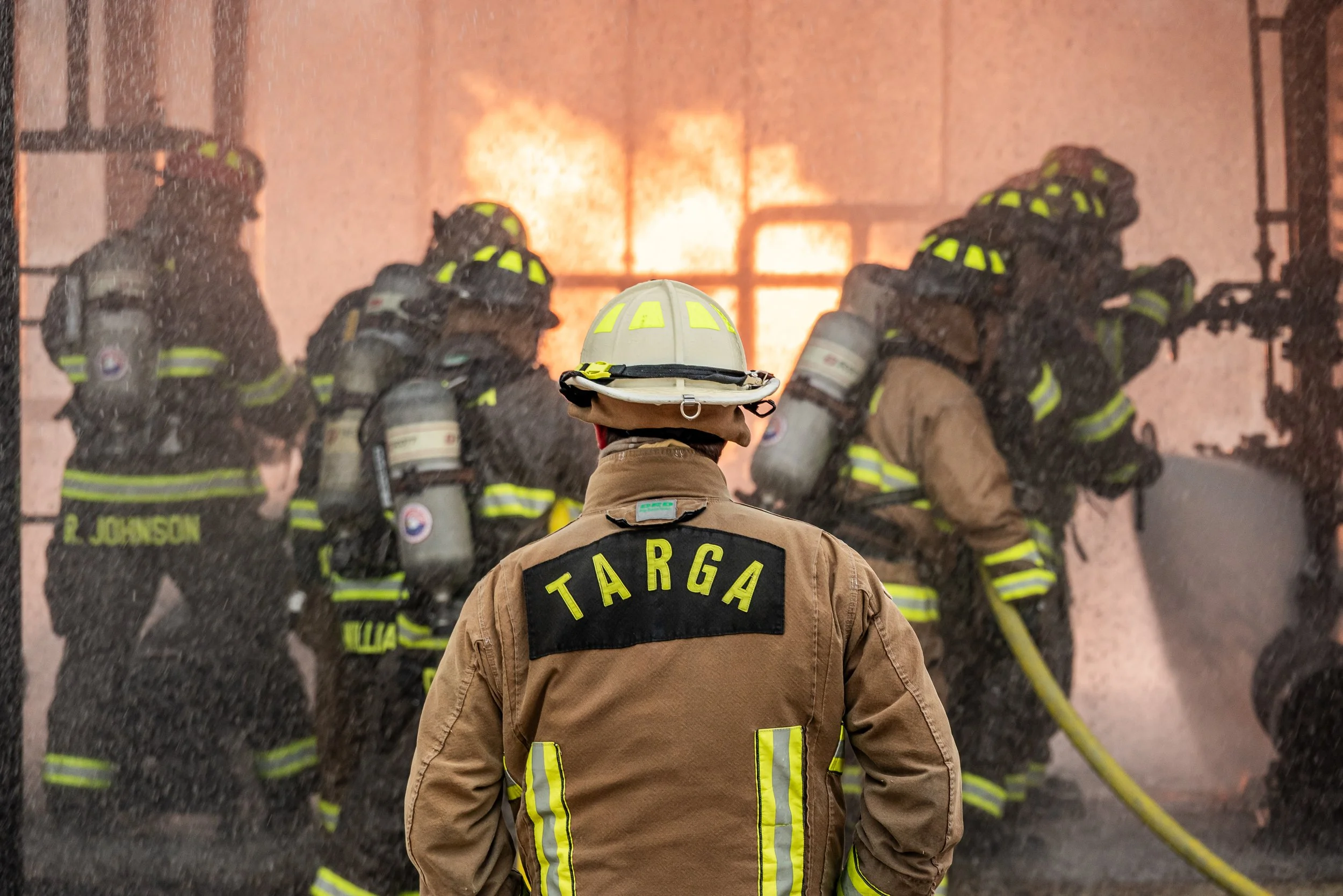 Firefighters in protective gear battling a blaze with flames and smoke in the background, viewed from behind a firefighter with 'Targa' on their uniform.
