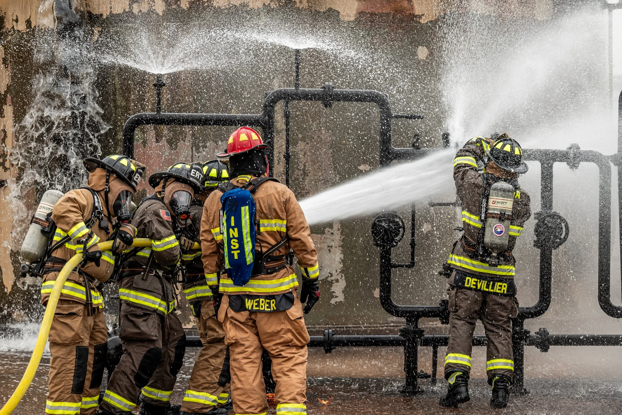 Group of firefighters in protective gear working together to spray water on a fire or fire-related incident at a facility with black pipes in the background.