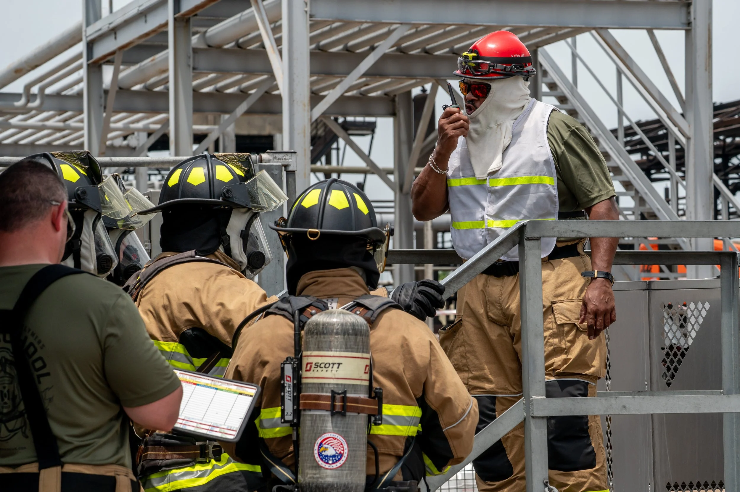 Firefighters and an emergency responder on a construction site, with some wearing protective gear and helmets, listening to a supervisor.