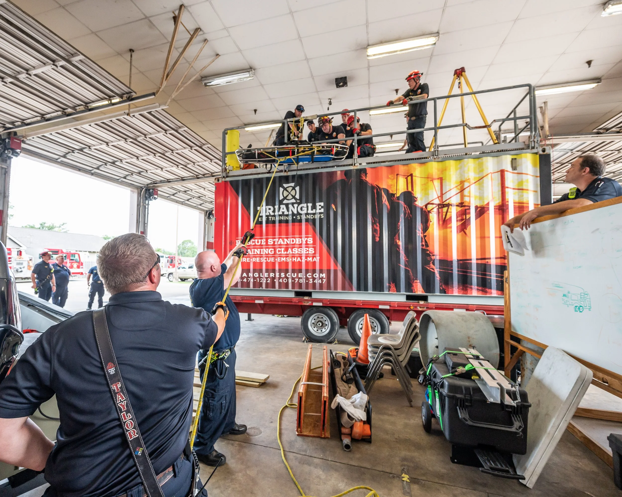Firefighters training in a garage, practicing rescue techniques using a harness and rope. People are observing and supervising.