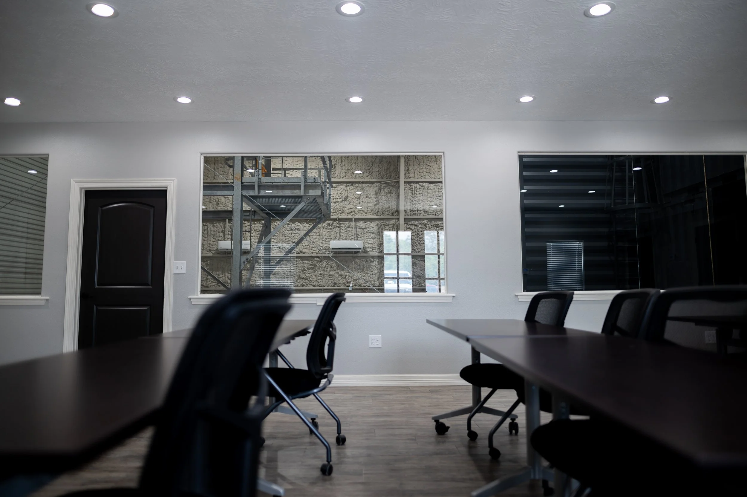 Empty conference room with black chairs, dark brown tables, a closed black door, and large windows showing scaffolding outside.