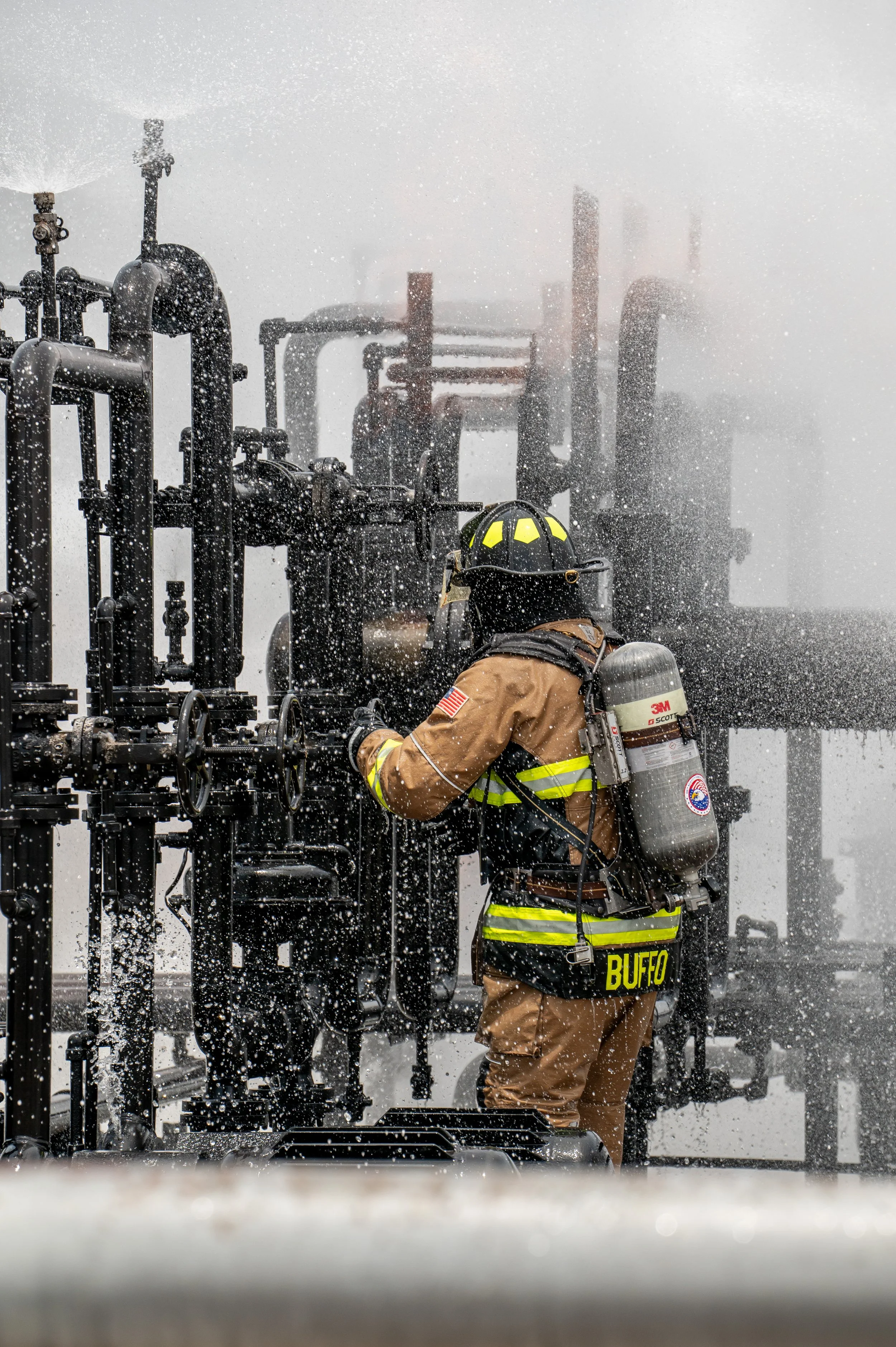 A firefighter in full gear working on industrial equipment amid heavy smoke and water spray.