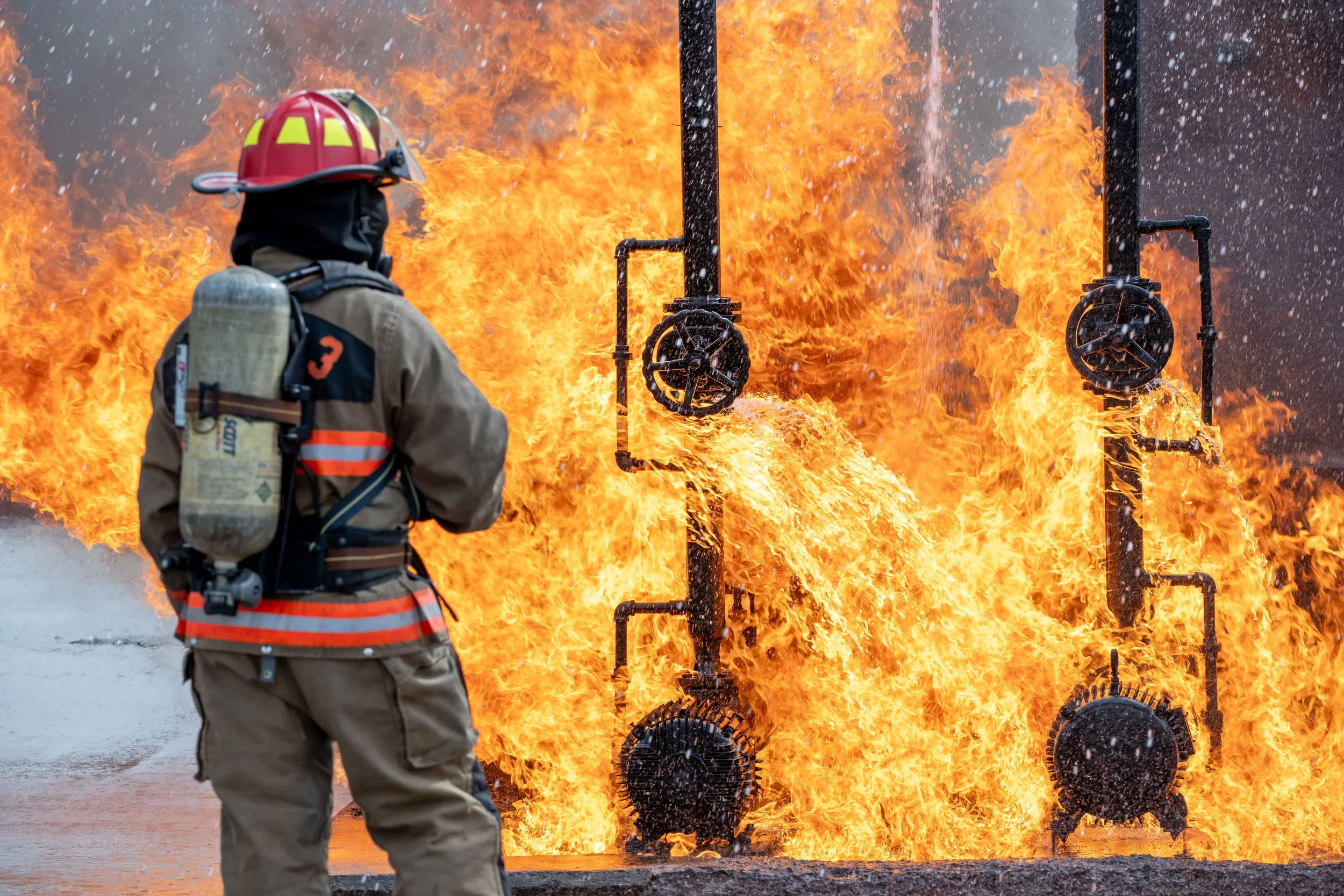 A firefighter in protective gear and a helmet standing with their back to the camera, facing a large fire and burning pipes.
