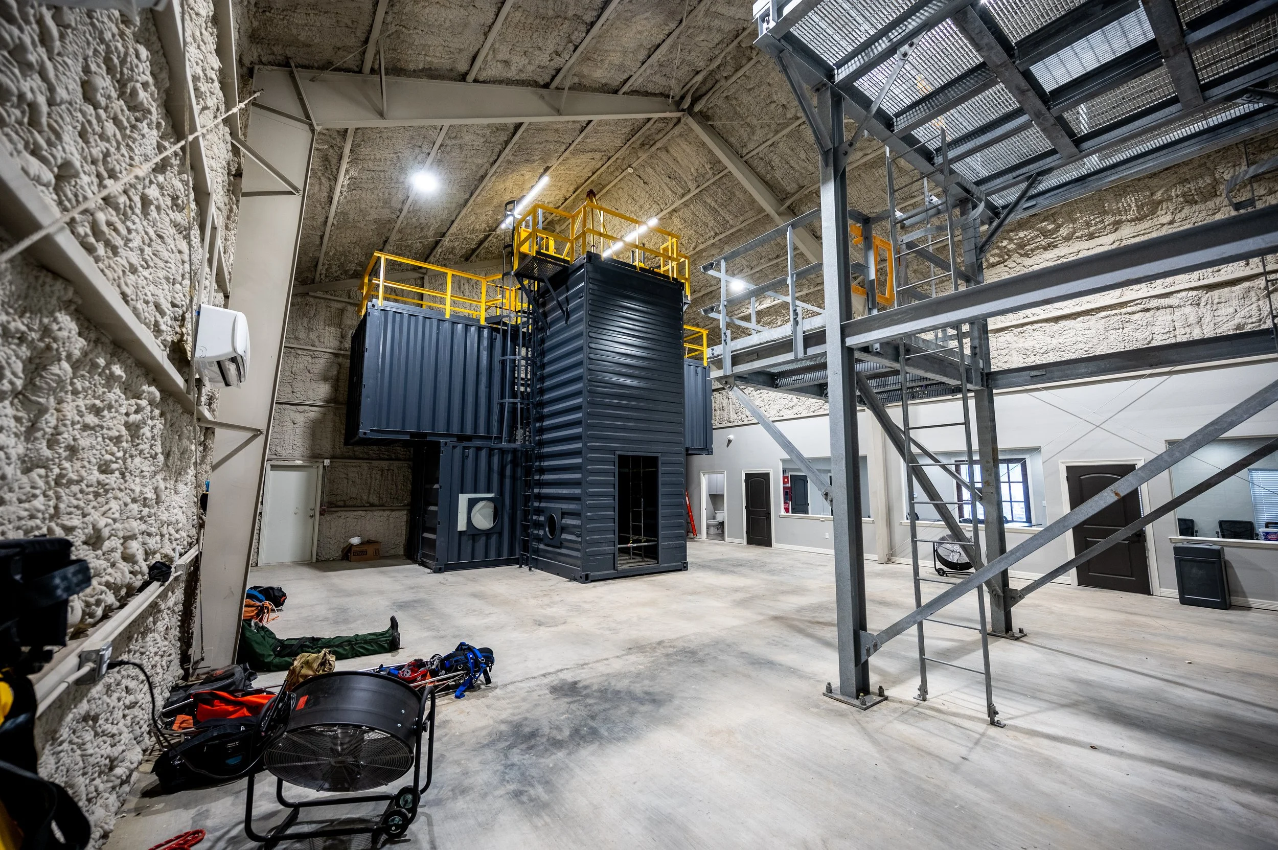 Interior of a large industrial or storage space with metal staircases, platforms, and two large black shipping containers.