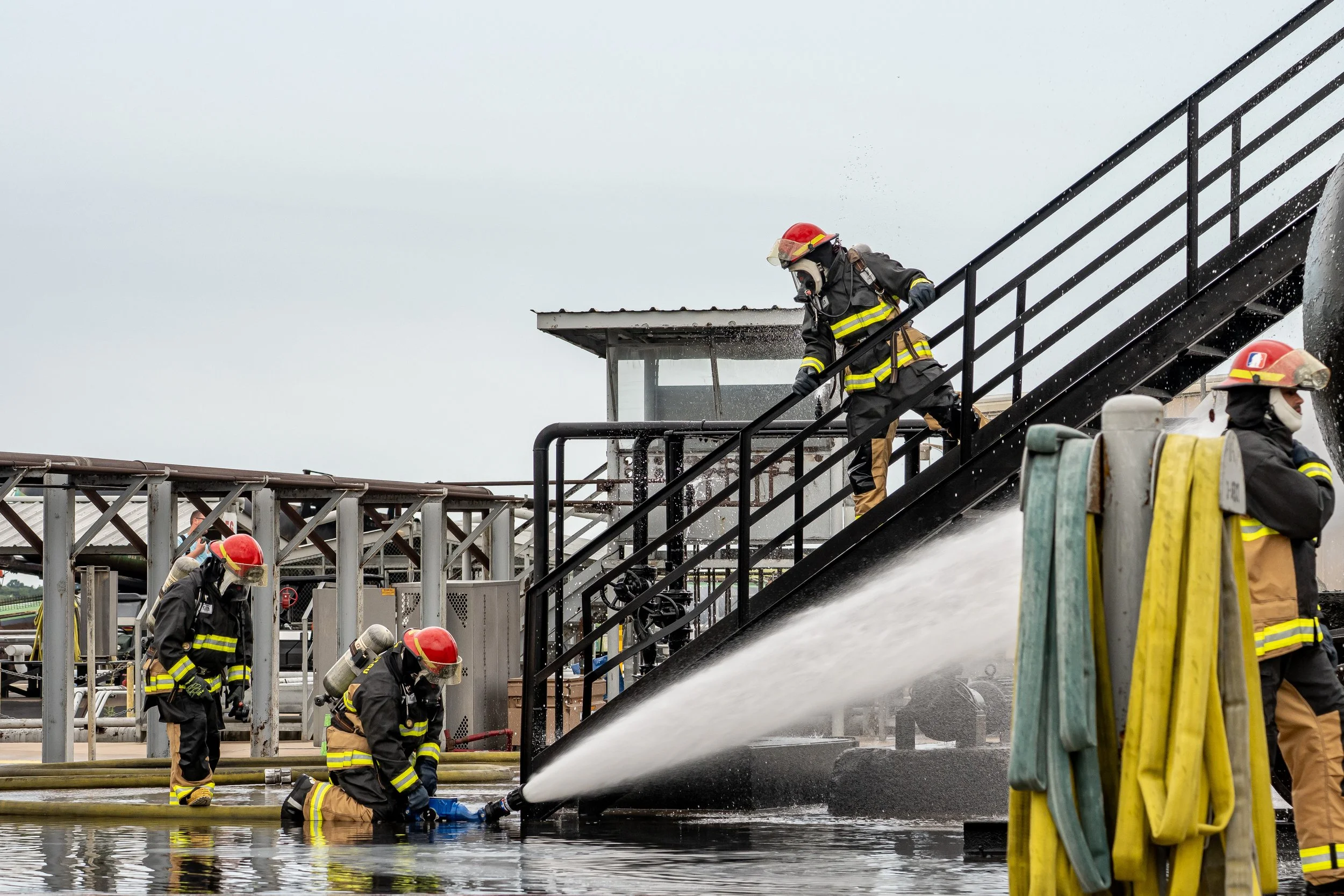 Firefighters working to spray water on an emergency situation at an industrial site, with some firefighters on the stairs and others on the ground, all wearing protective gear and helmets.