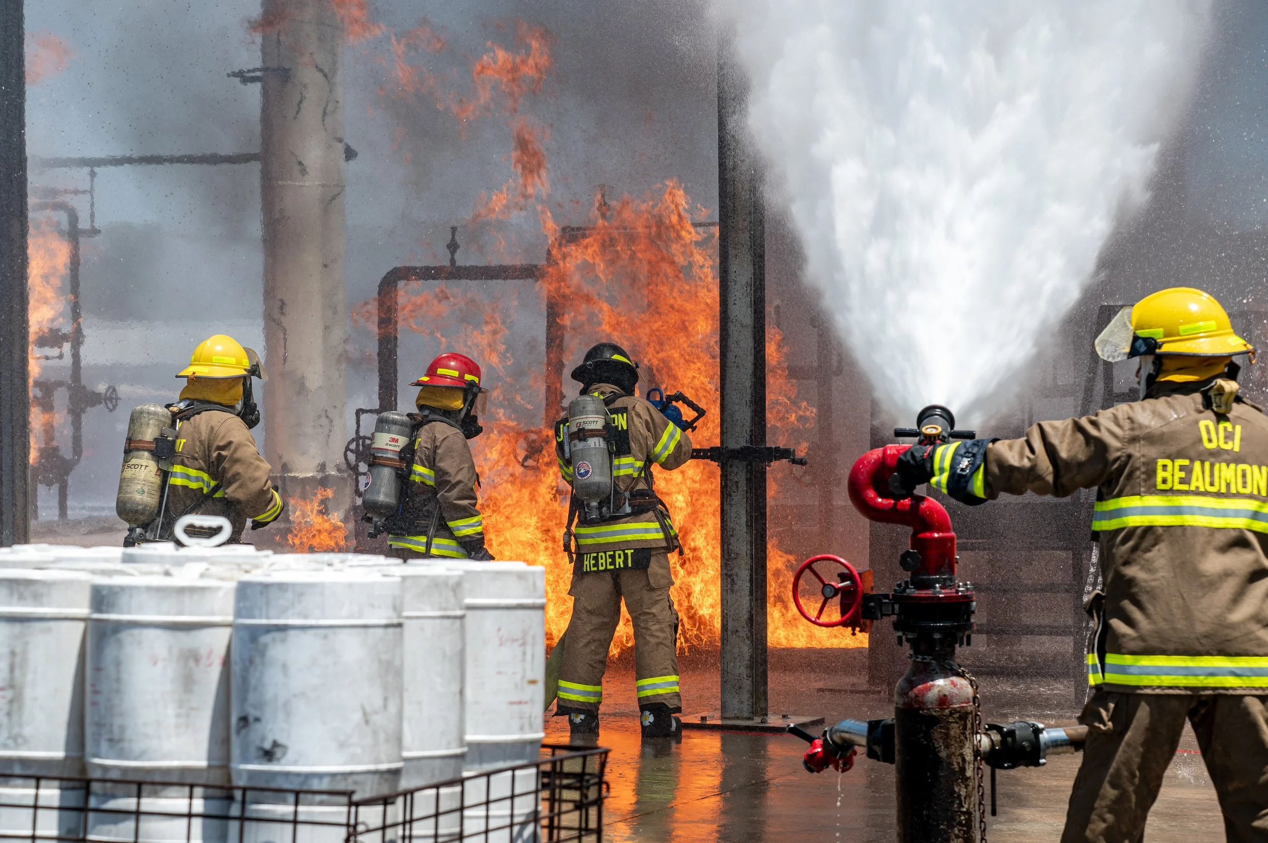 Four firefighters combat a large industrial fire at night, with flames and smoke rising in the background. They are wearing protective gear and helmets, with some using hoses to spray water on the fire.