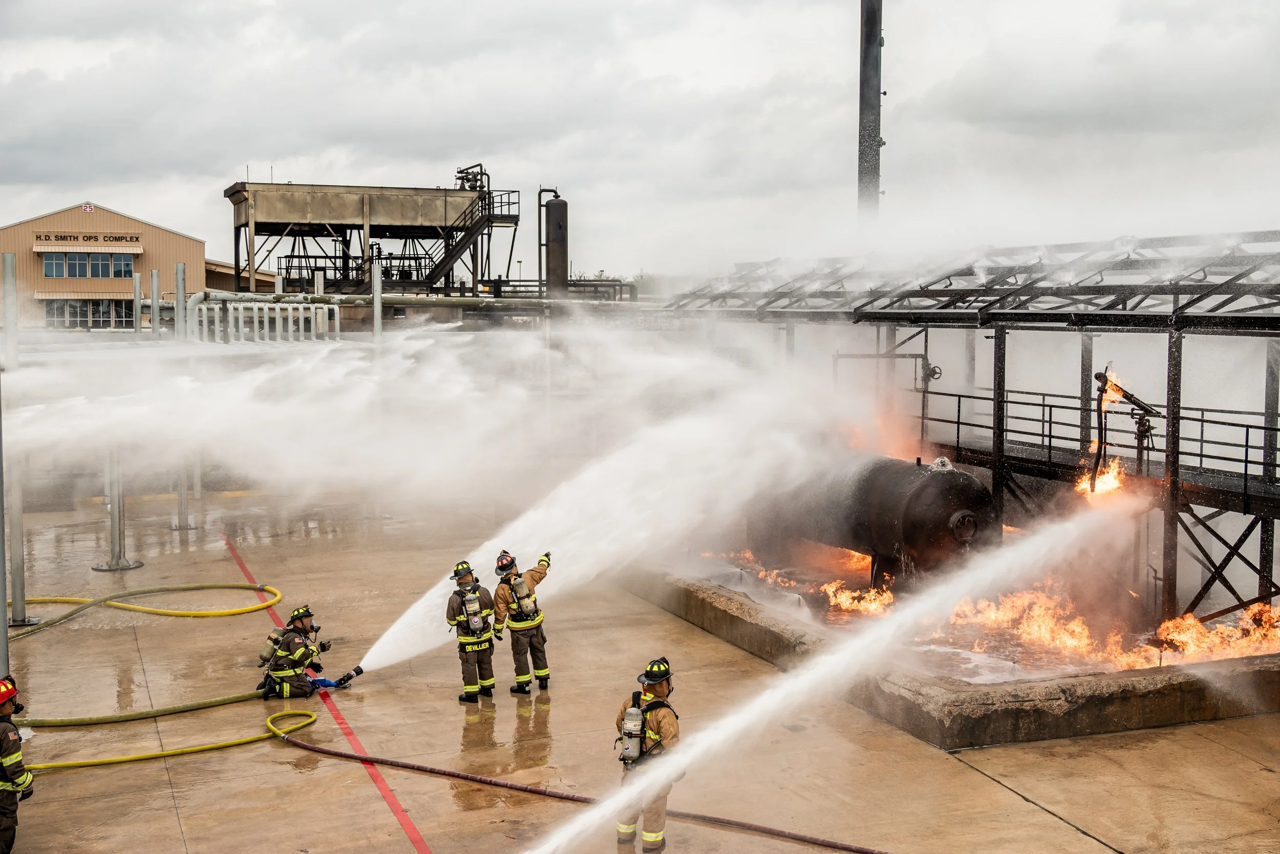 Firefighters fighting a fire with hoses in an industrial area with flames and smoke, a large cylindrical tank on fire, and industrial structures in the background.