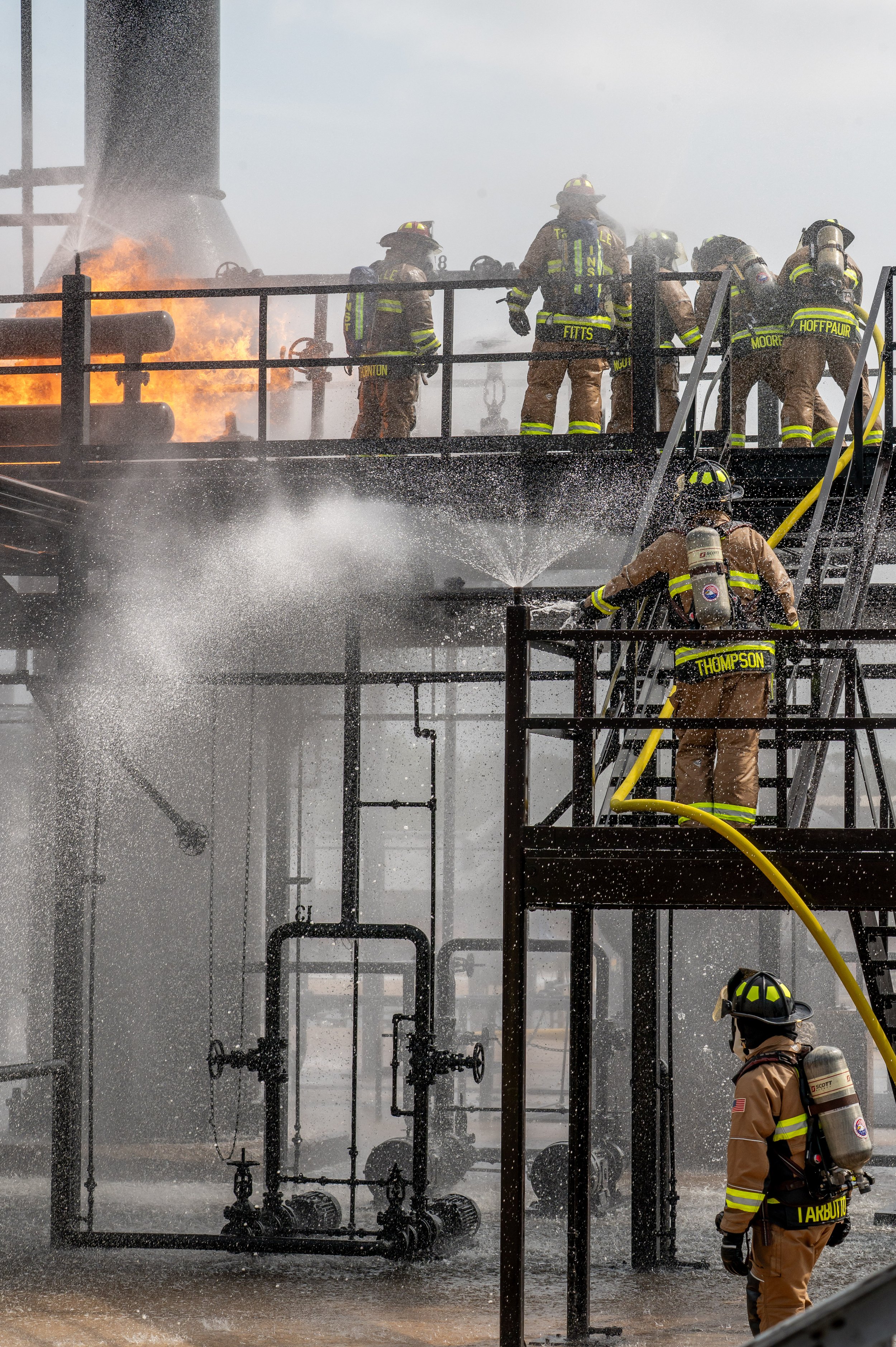 Firefighters in firefighting gear spraying water on a fire on an industrial or refinery structure, with smoke and flames visible, and metal pipes and ladders around them.