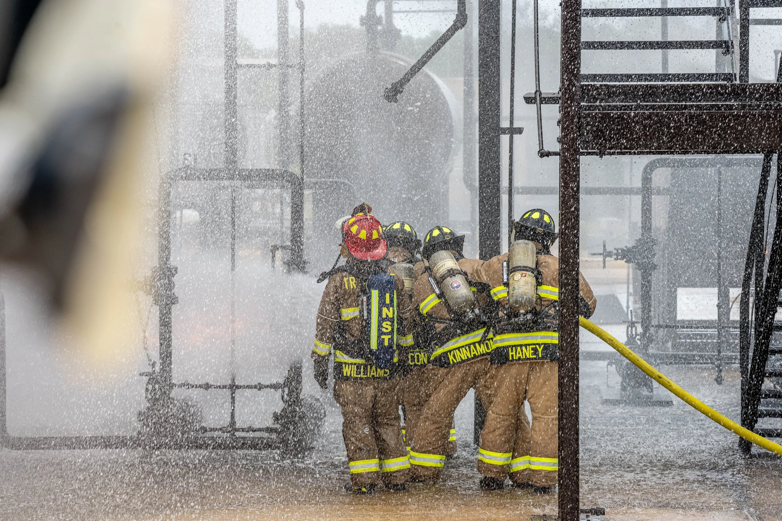 Four firefighters in full gear work to contain a fire during a heavy rainstorm, with water spraying around them.