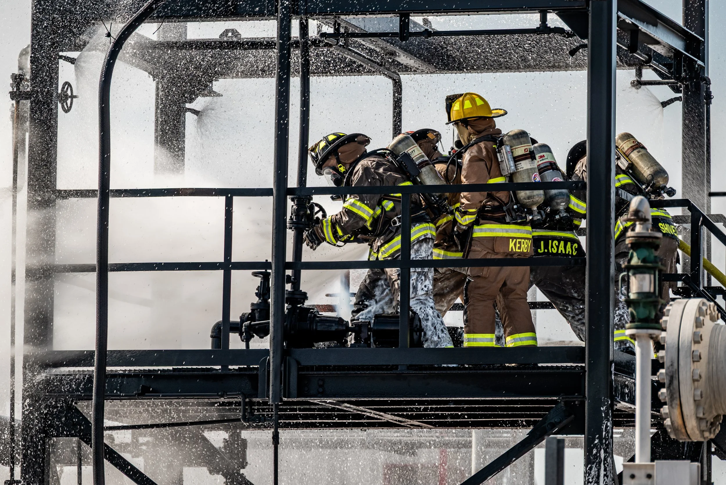 Firefighters wearing protective gear and helmets working on upper platform of industrial fire scene, spraying water from hoses amidst water spray and smoke or steam.
