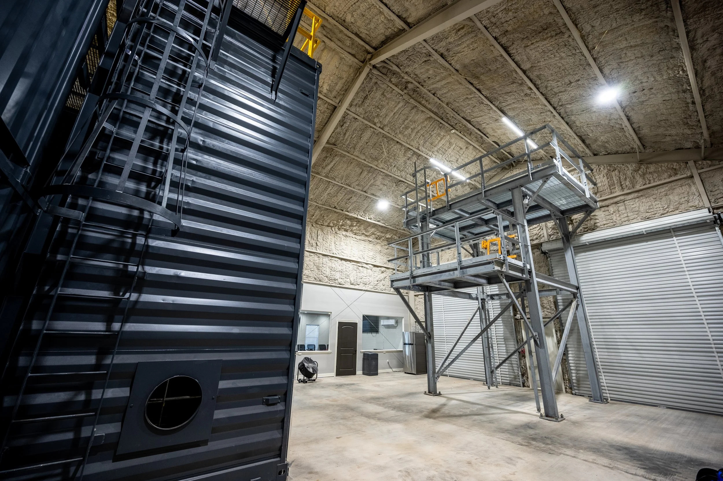 Empty industrial warehouse with high ceiling, metal staircase, and large metal container on the left side.