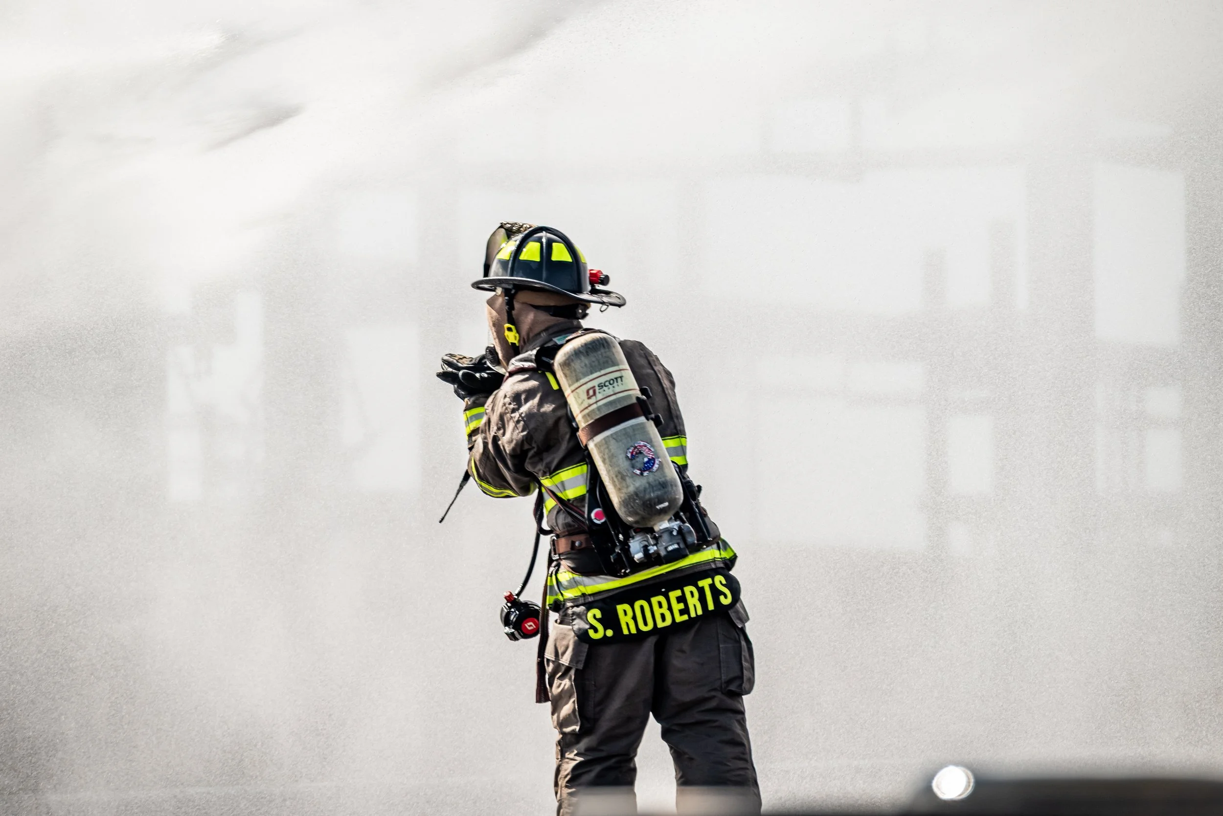 Firefighter in full gear, including helmet, reflective stripes, and a breathing apparatus, standing outdoors on a cloudy day.