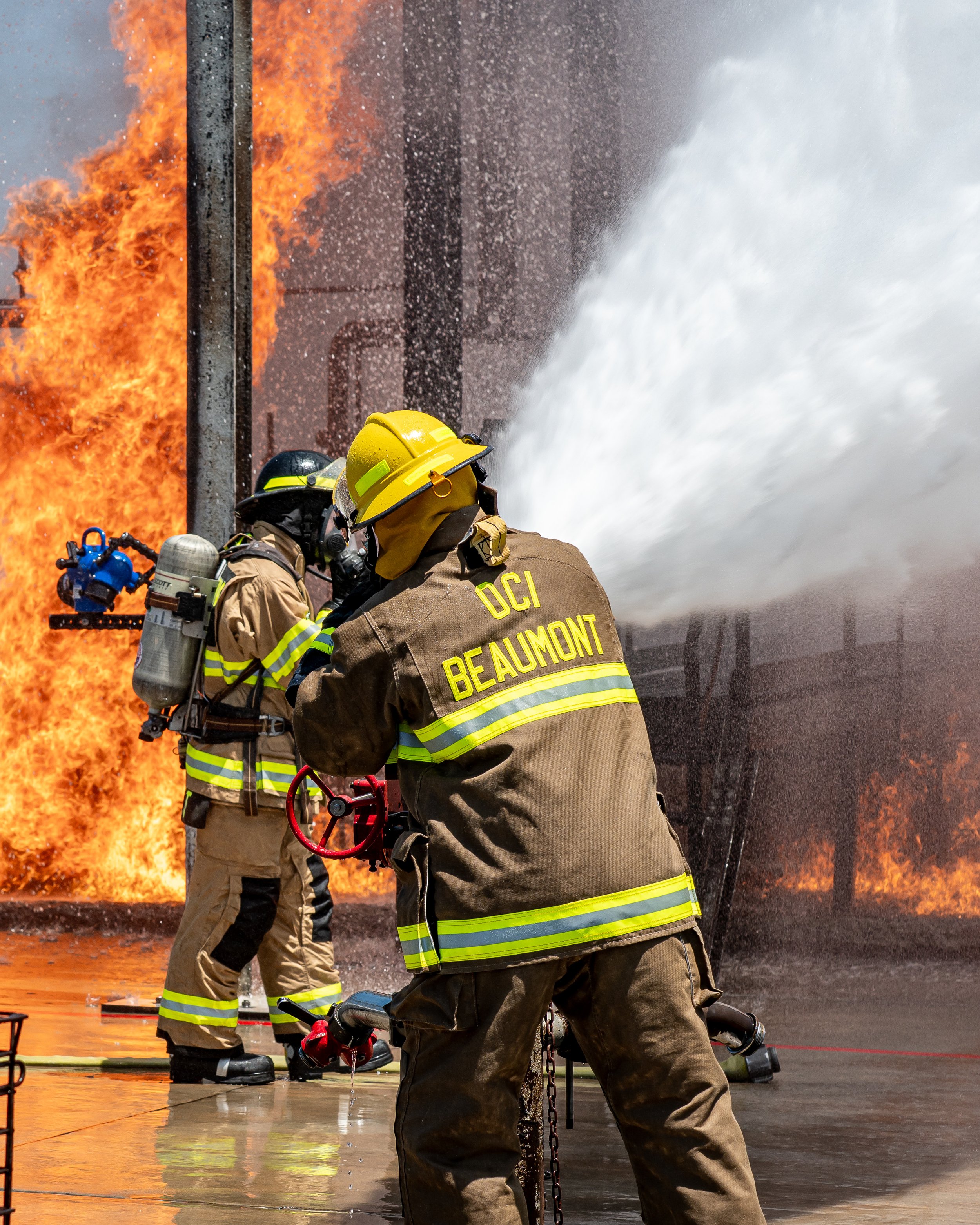 Firefighters fighting a large fire with water hose, flames and thick smoke in the background, wearing firefighting gear with 'OC' and 'Beaumont' on their jackets.