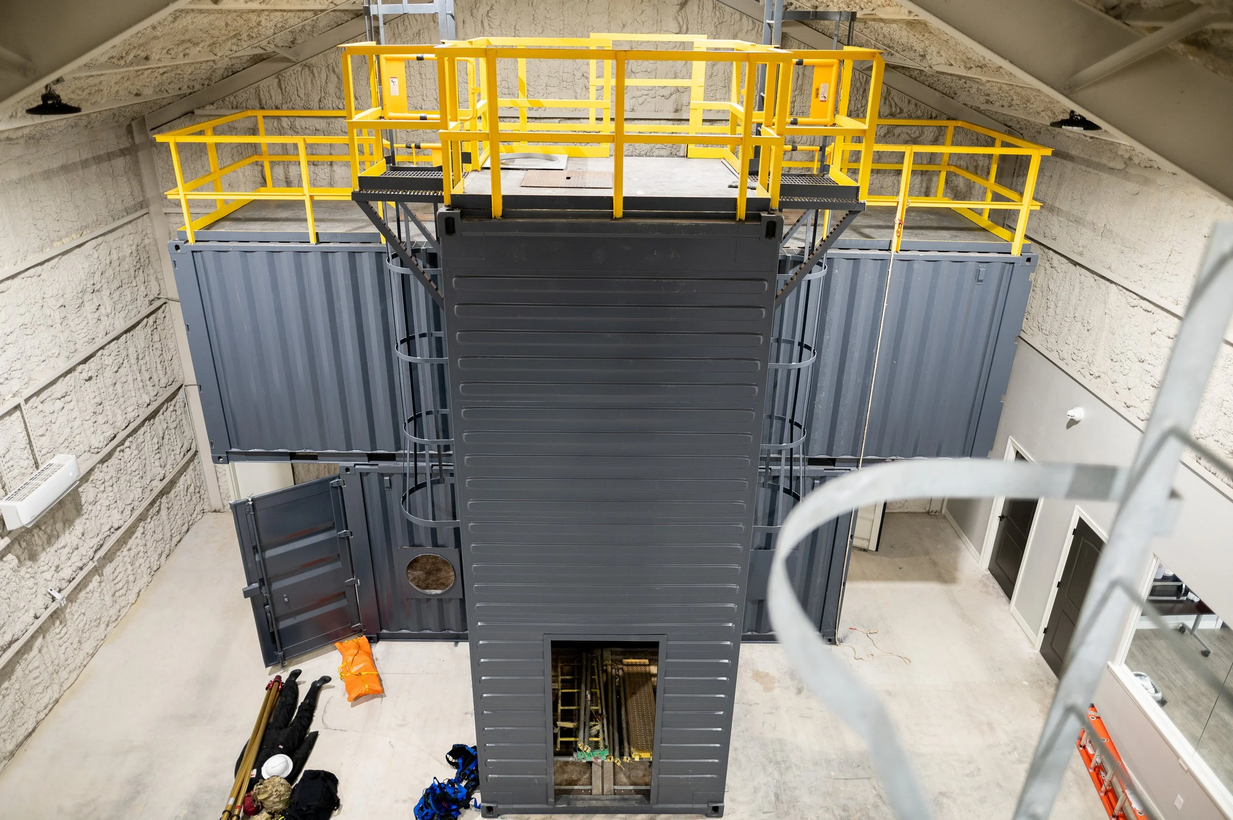 Construction site of a two-story structure made from modular shipping containers, with a staircase leading to a rooftop platform with yellow safety railings, inside a warehouse or industrial building.