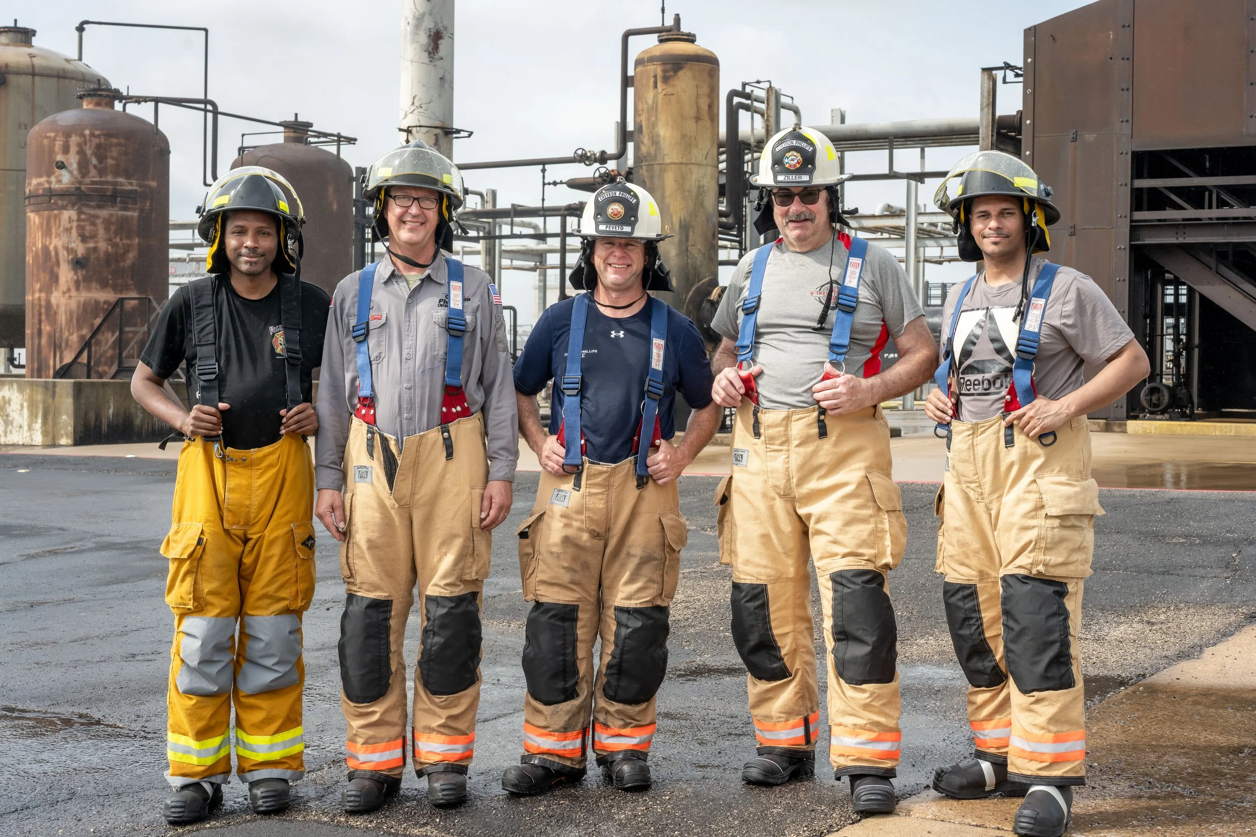 Six firefighters in protective gear standing in front of an industrial facility, smiling at the camera.