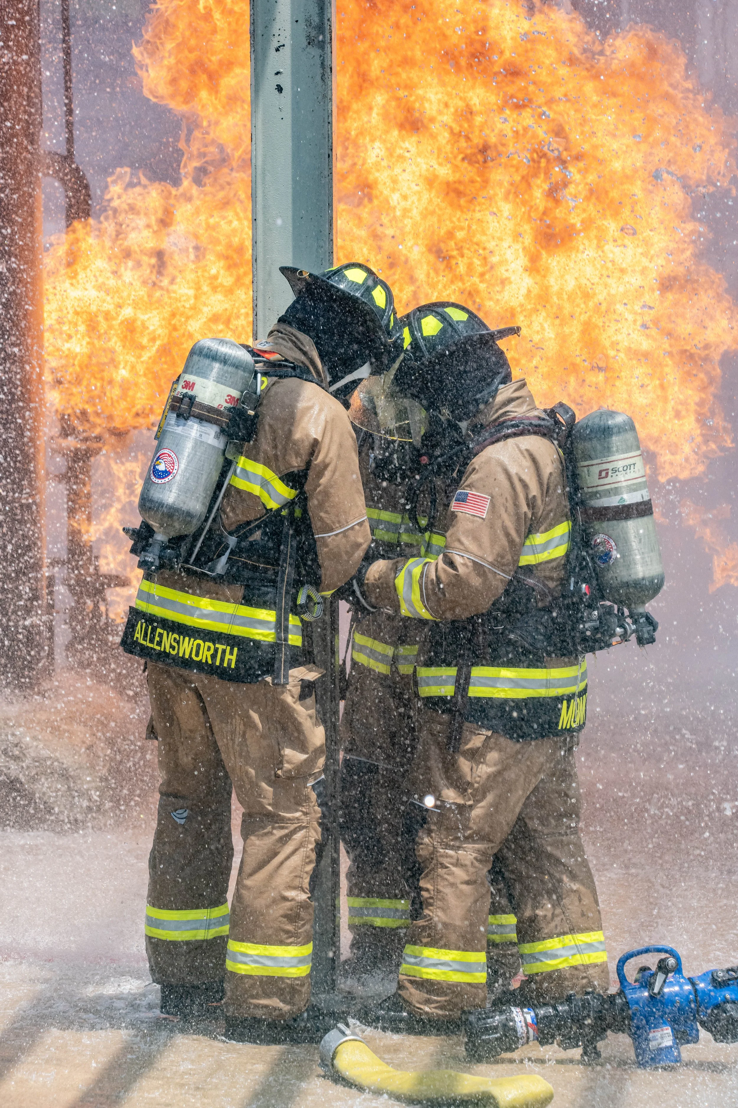 Three firefighters in full gear fighting a large blaze with intense flames and smoke in the background.