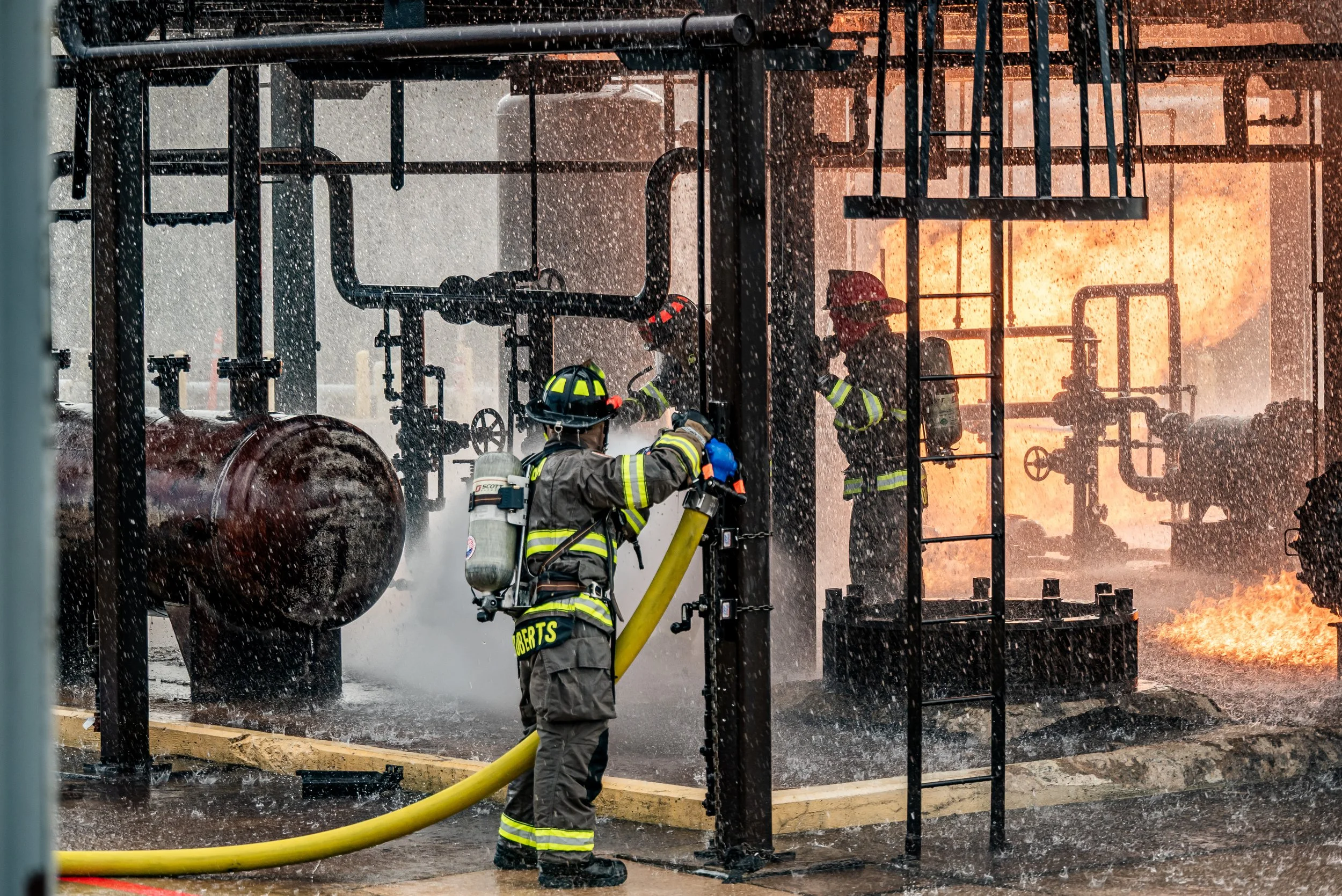 Firefighters in protective gear combating a fire at an industrial facility, with flames visible in the background and water spraying onto the flames.