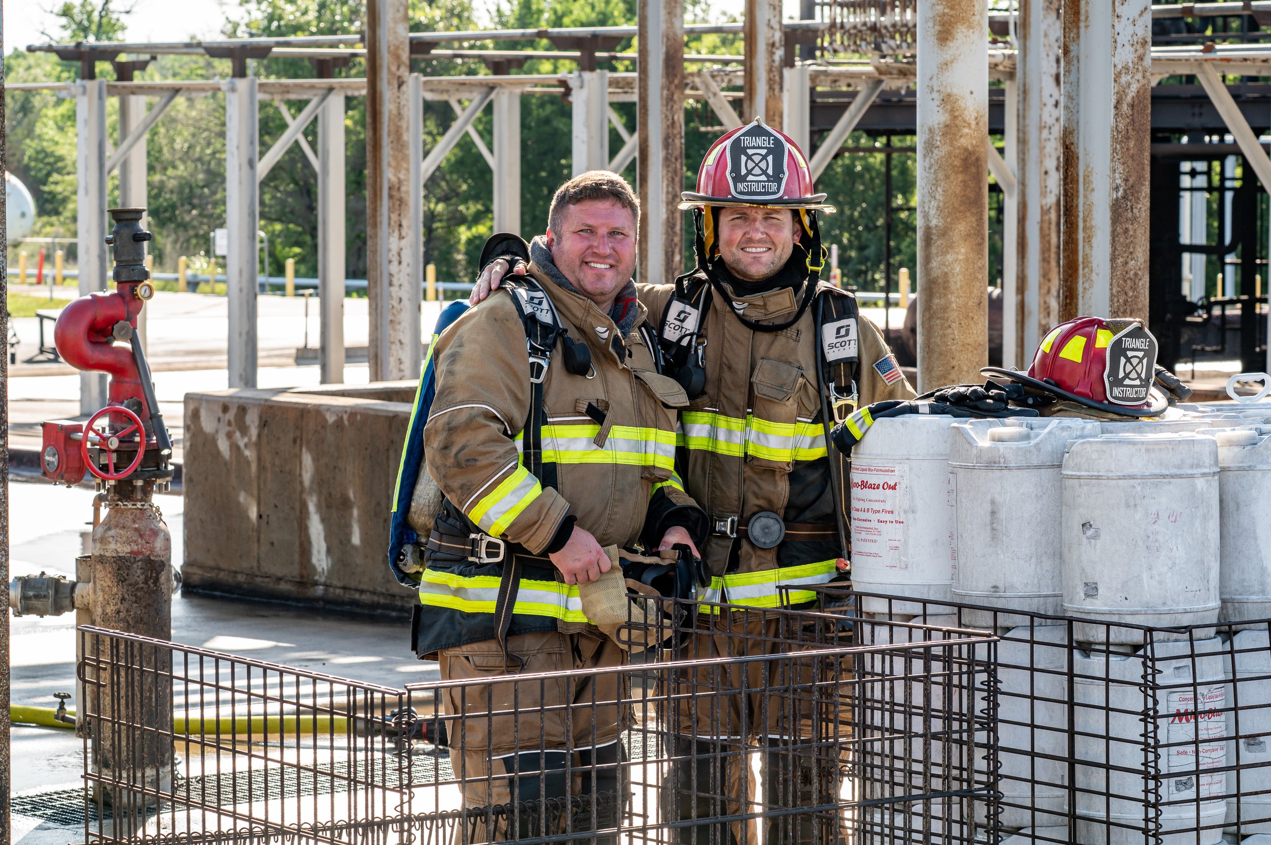 Two male firefighters in full gear smiling and standing side by side outdoors at a fire station or training facility, with equipment and a large metal container nearby.