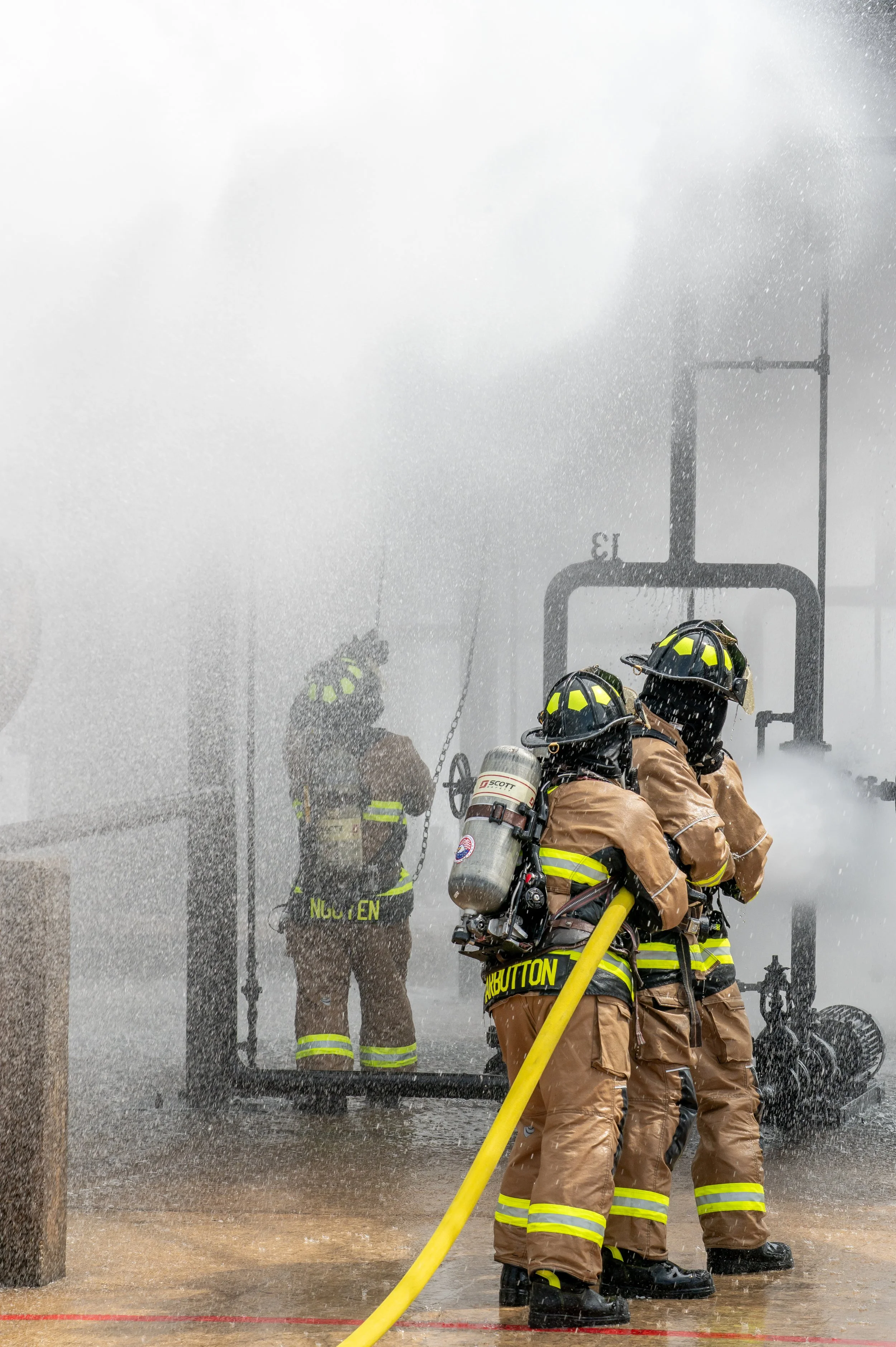Firefighters wearing full gear and helmets, with one operating a fire hose, battling a fire with water spray and smoke.