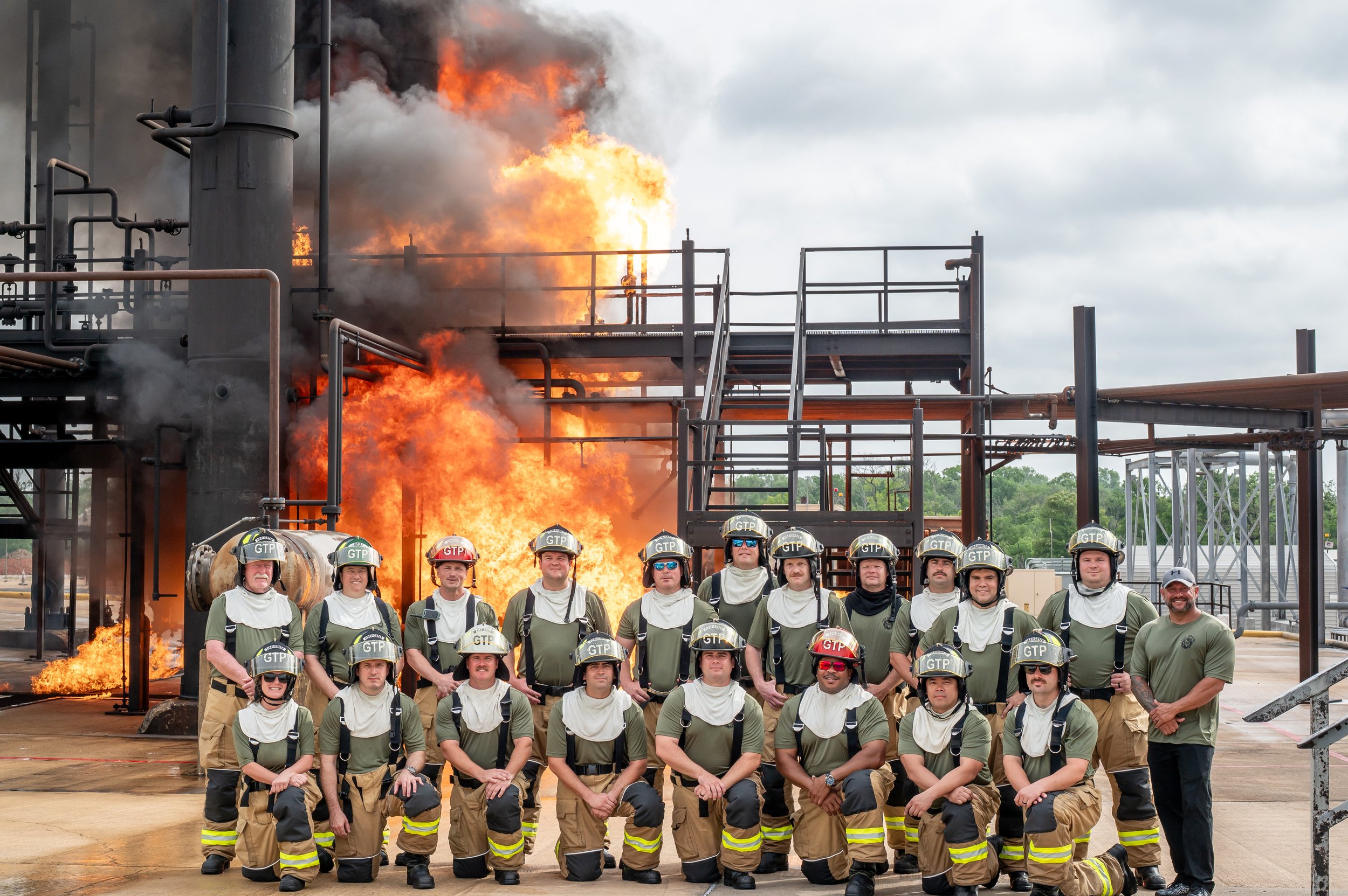 Group of firefighters in front of a large industrial fire with flames and smoke, posing for a photo.