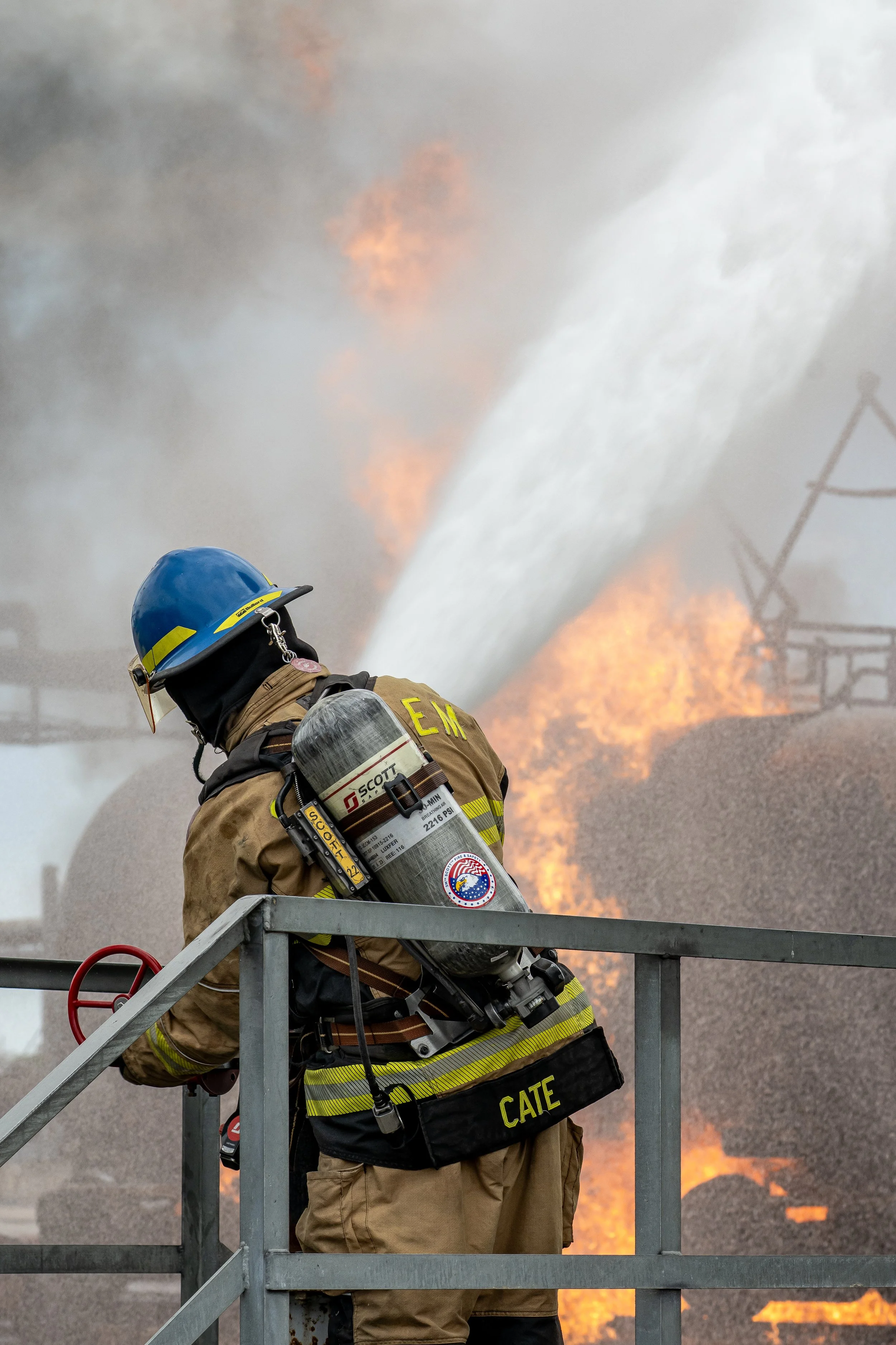 Firefighter in full gear, wearing a blue helmet and an air tank, stands on a platform during a large fire with flames and thick smoke in the background.
