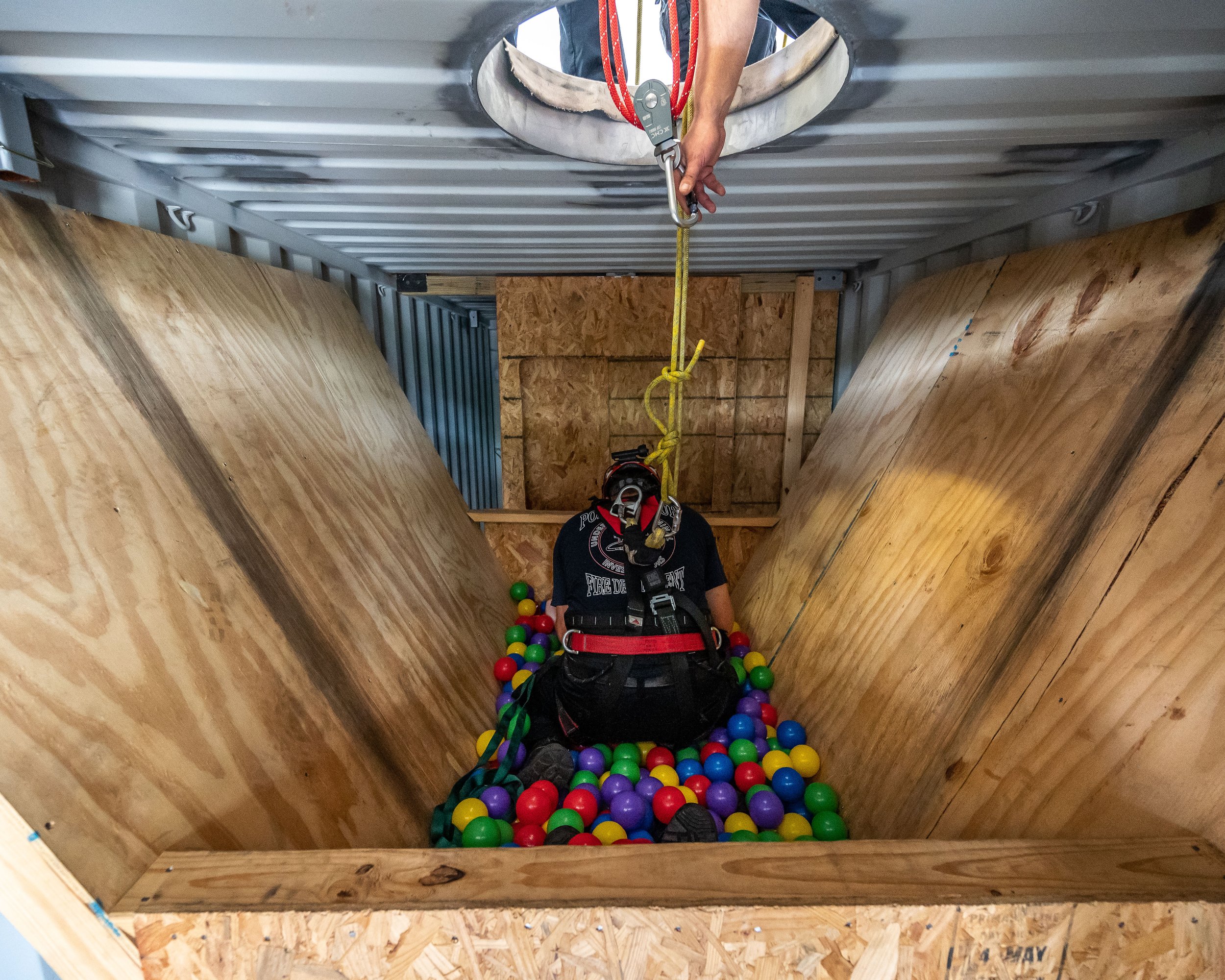 A person in safety gear and a helmet descending into a wooden ball pit with colorful plastic balls, using a harness attached to a pulley system.