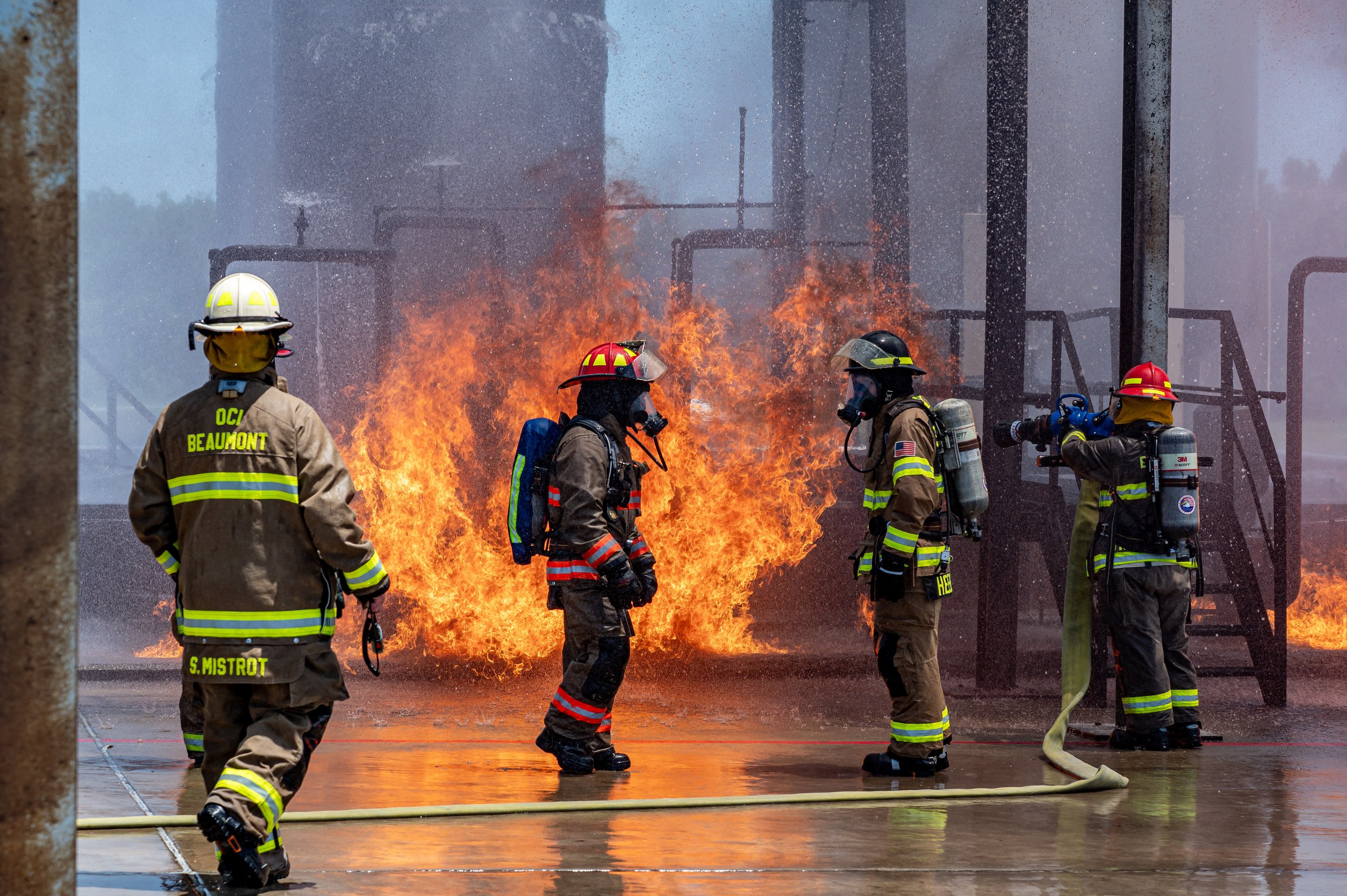 Firefighters in full gear working to extinguish a fire with large flames and water spray at an industrial site.