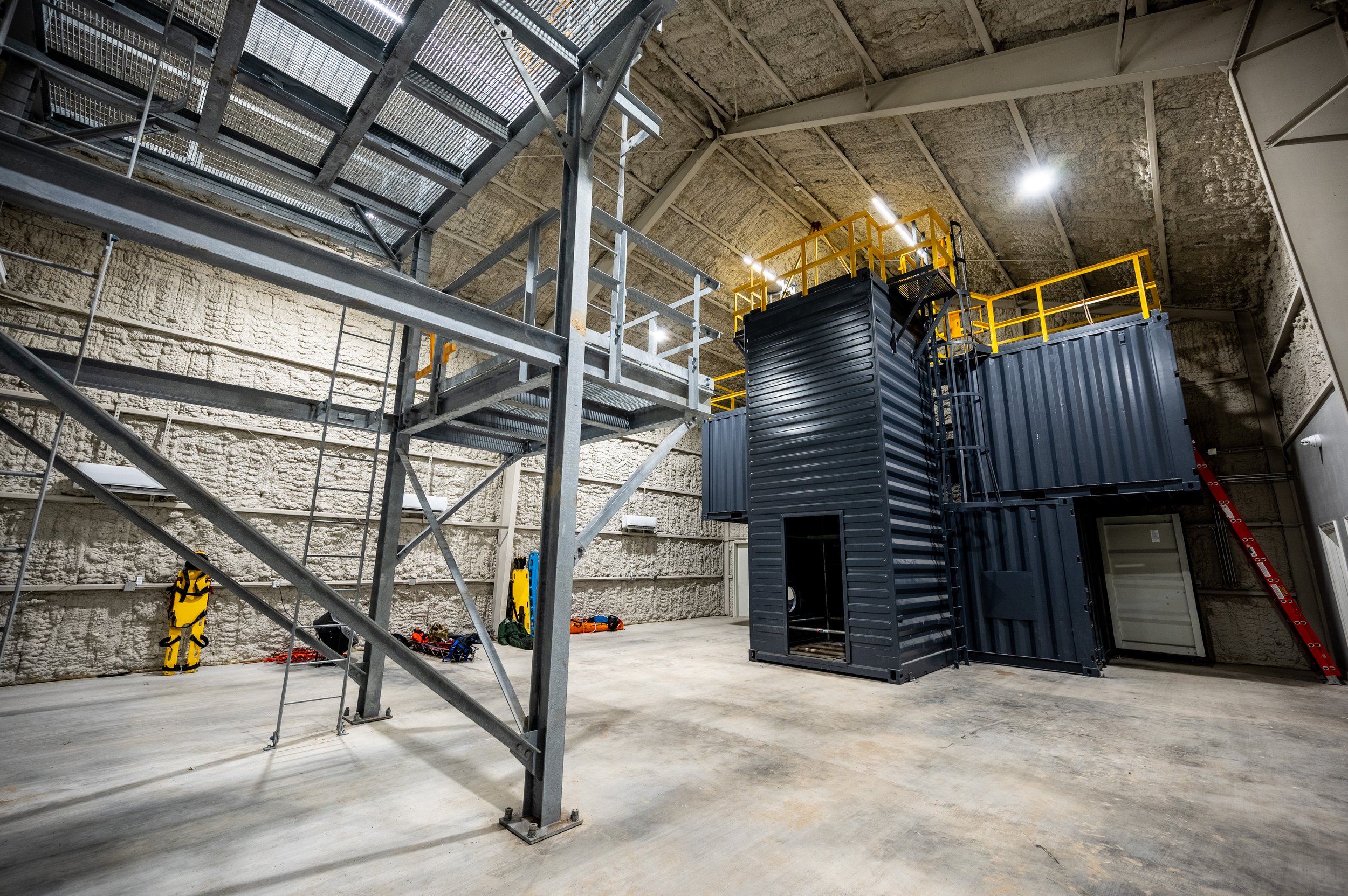Empty industrial or scientific space with metal stairs and platforms, and black metal containers or structures, in a high-ceiling room with textured insulation walls.
