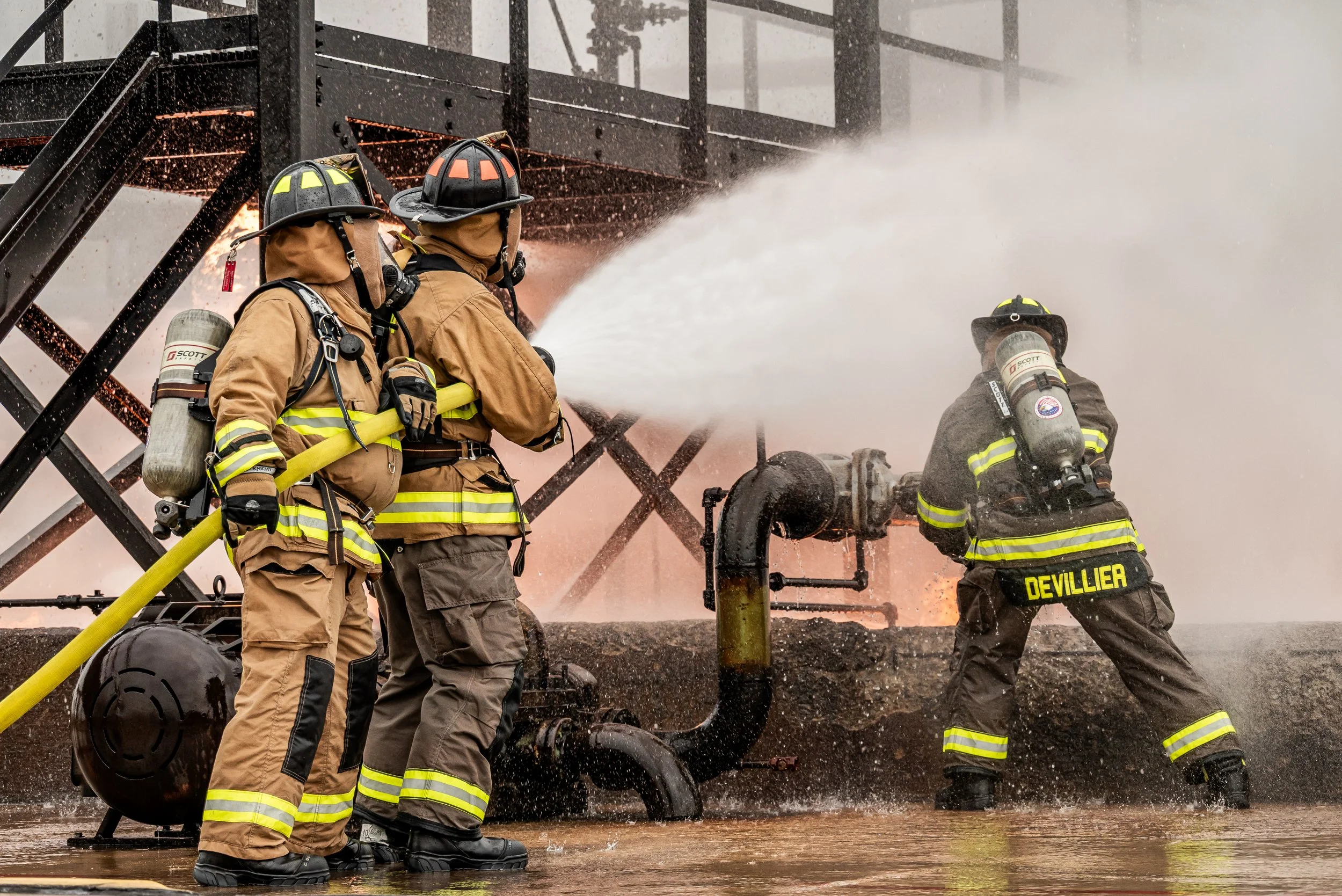 Three firefighters in full gear fight a fire with a hose, with water spraying and steam rising, near a metal structure.