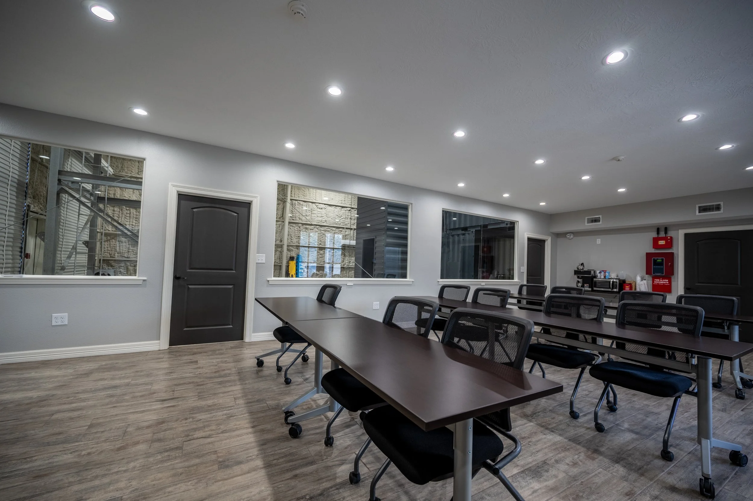 Empty conference room with front-facing tables, chairs, windows, and a fire alarm and safety equipment on the back wall.