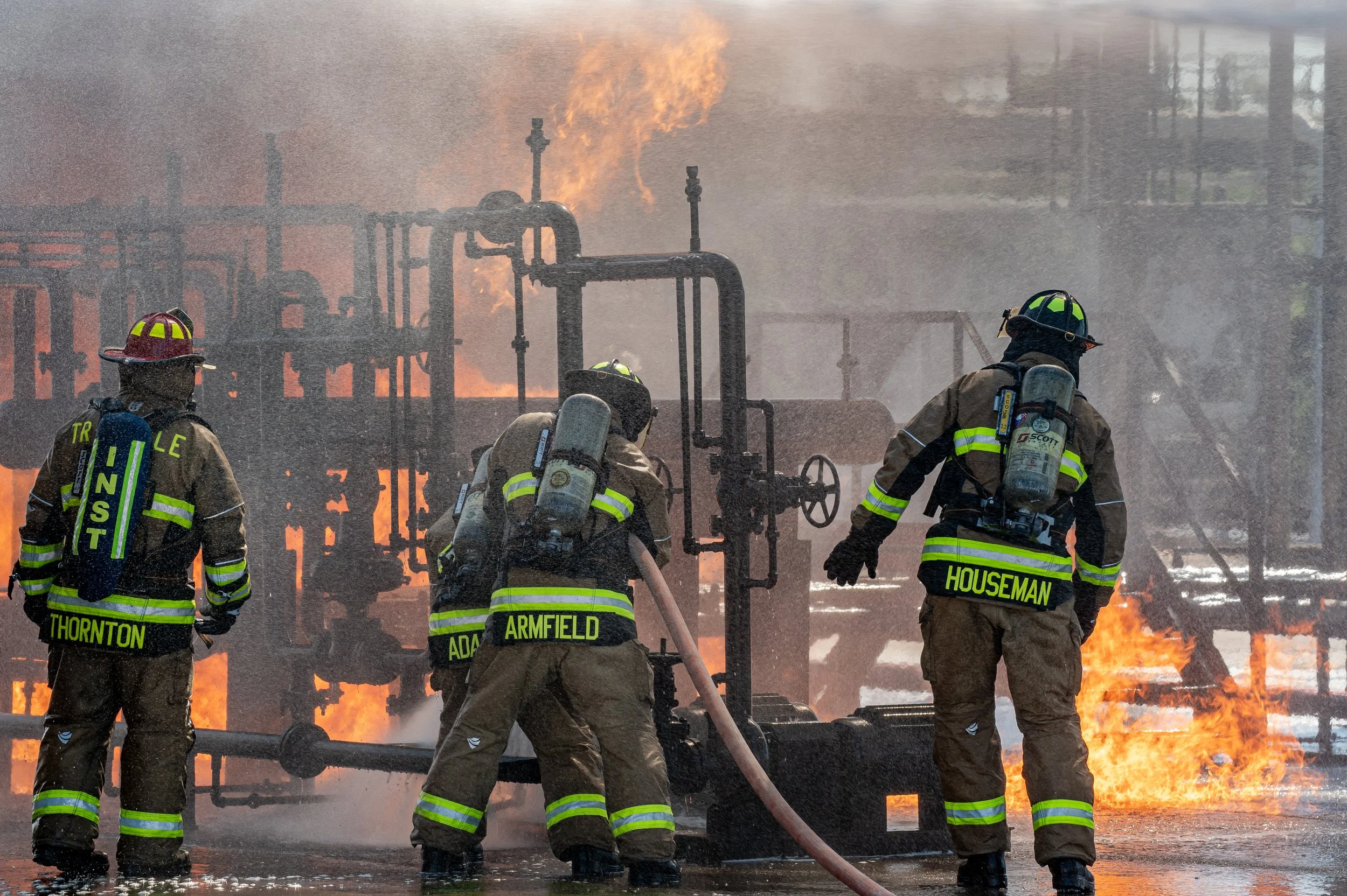 Four firefighters in protective gear are fighting a fire at an industrial site, with flames and smoke visible in the background.