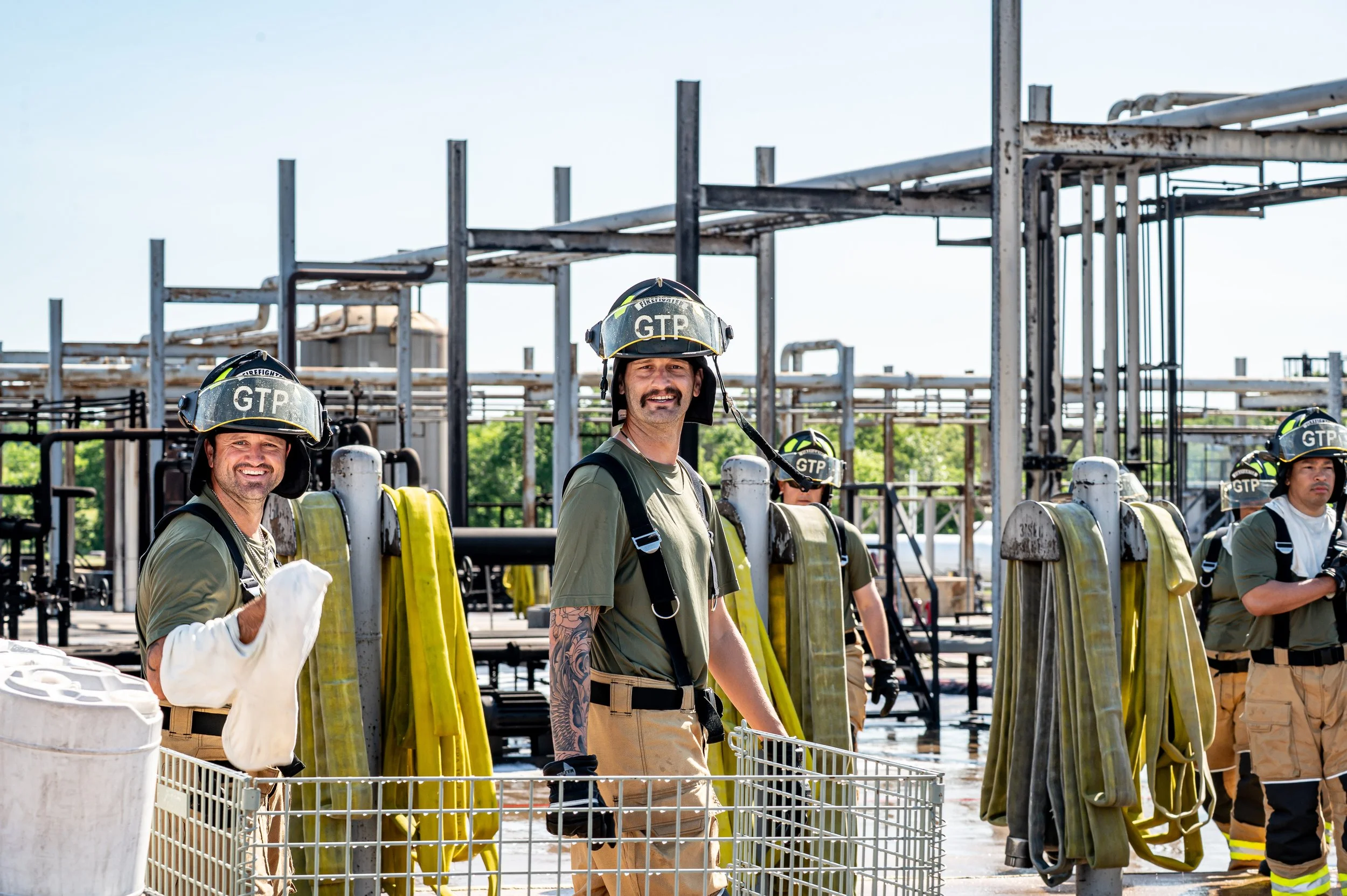 Firefighters in gear and helmets standing at a fire station. They are smiling and adjacent to equipment and hoses, with a metal framework structure in the background.