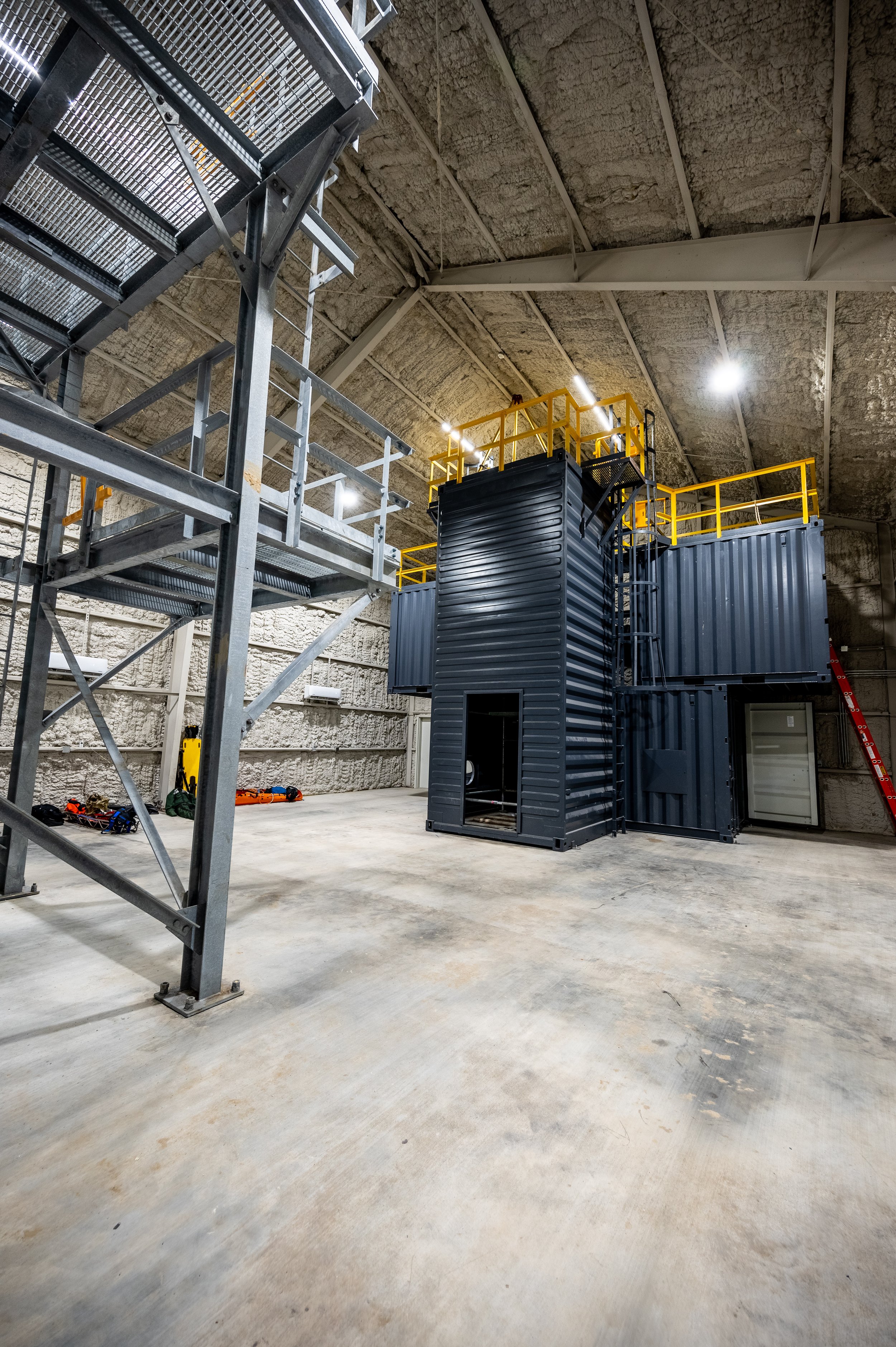 Interior of an industrial building with metal scaffolding, a black shipping container structure with yellow safety railings on the upper level, and a concrete floor and ceiling.