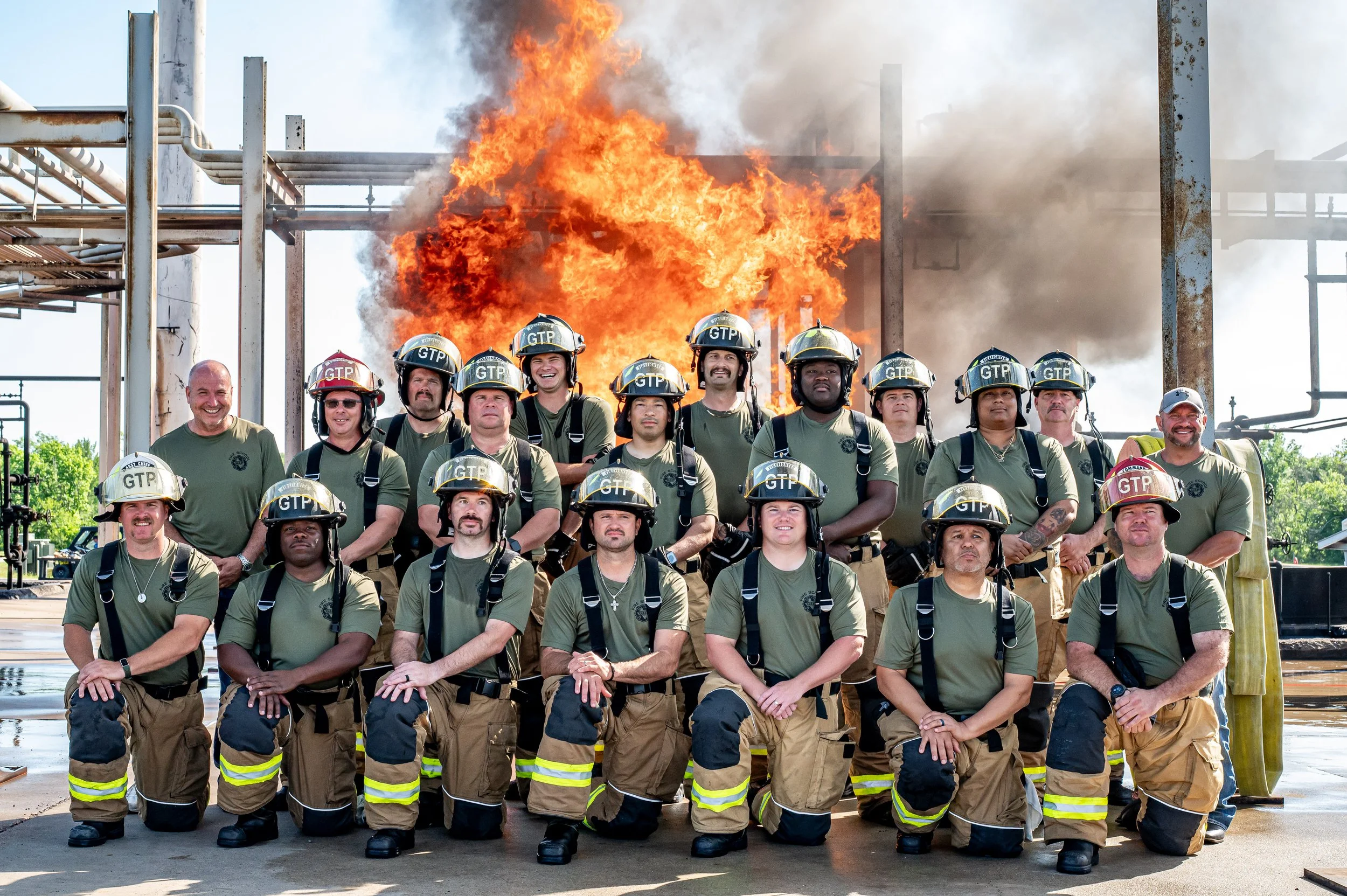Group of firefighters in uniform kneeling and standing in front of a large fire with flames and smoke at an industrial site.