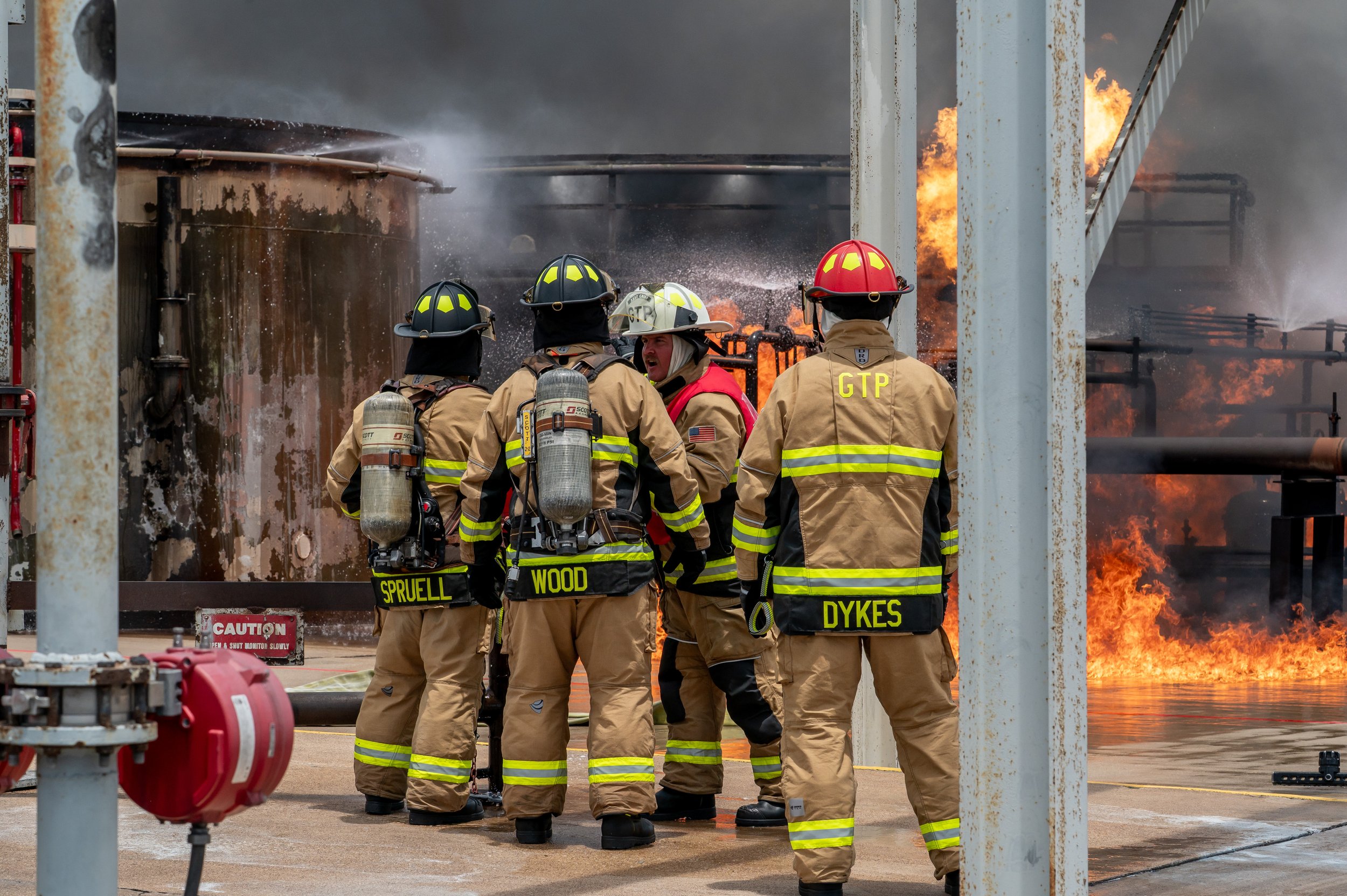 Firefighters in full gear, including helmets and oxygen tanks, gather in front of a large fire on an industrial site with flames and smoke in the background.
