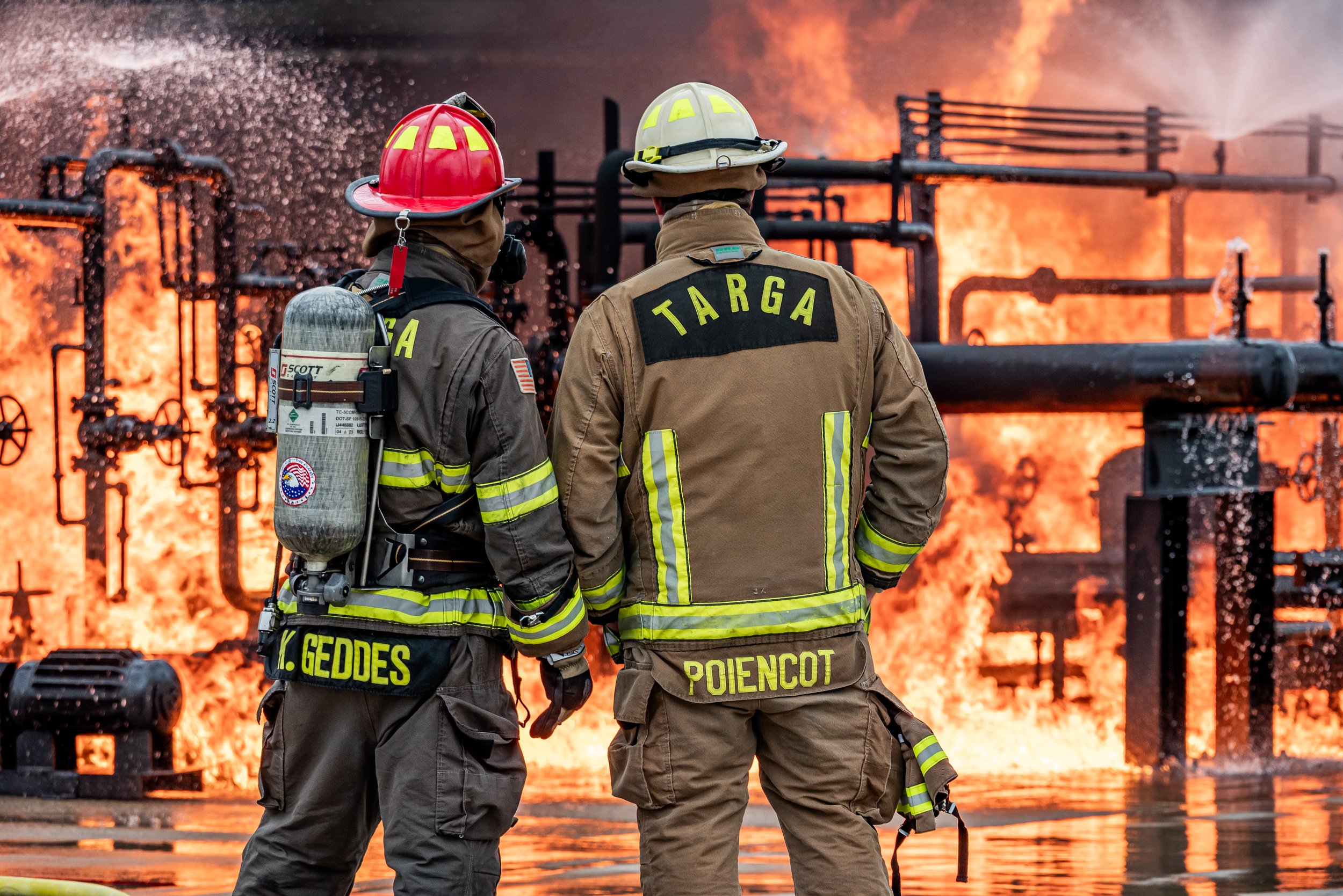 Two firefighters standing side by side in front of a large fire and flames, with industrial pipes in the background. One firefighter has a breathing mask on their back and the other has their hand on their hip. They are wearing protective gear and he