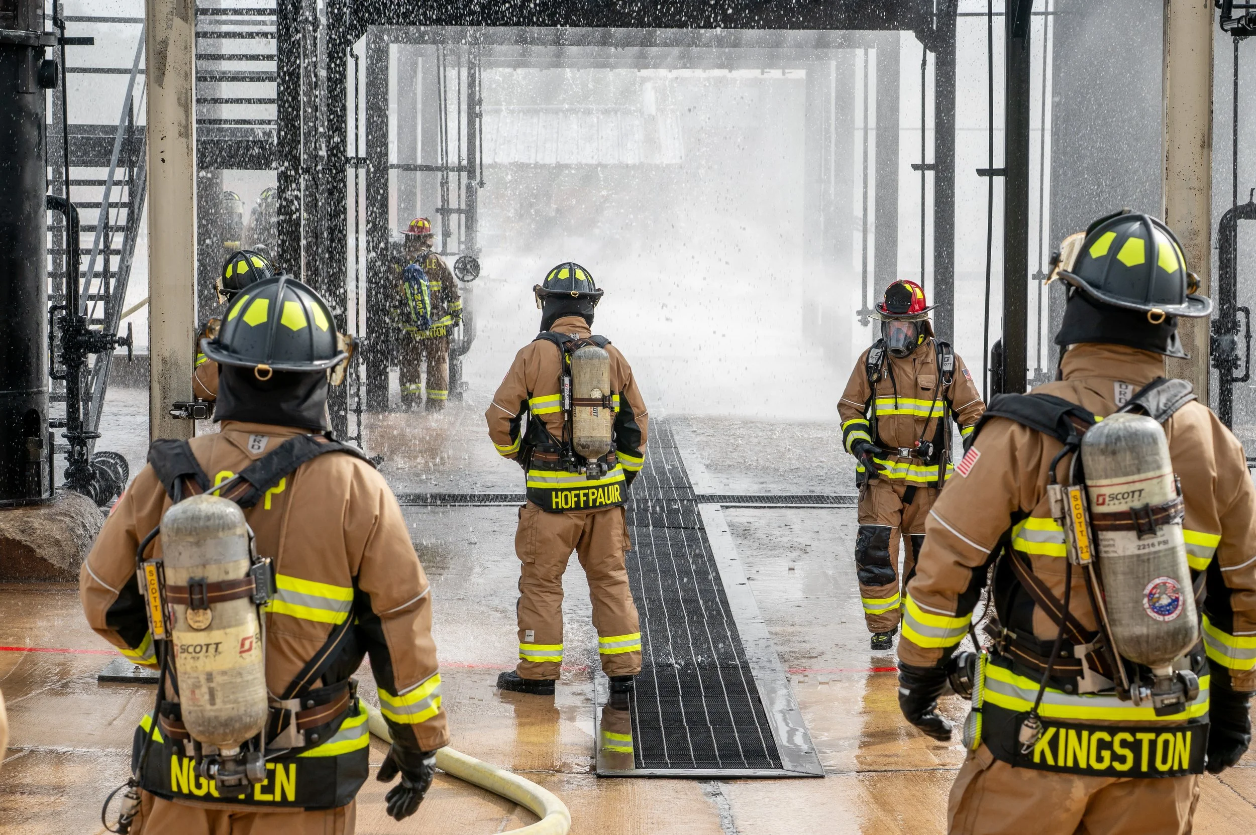Group of firefighters in protective gear fighting a fire at an industrial site with water spraying and steam visible.